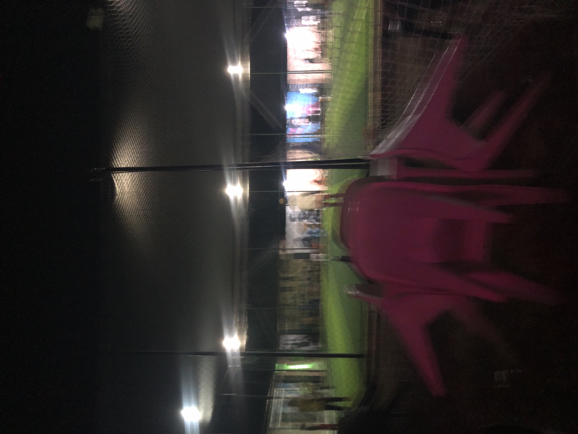 The image depicts an outdoor gathering at night, possibly in a recreational area or event venue in Pindiga, Nigeria. In the foreground, several bright pink plastic chairs are stacked or arranged, some of them out of focus due to motion blur. Behind the chairs, a green, well-lit field, likely artificial turf, is visible, suggesting a sports or event space. A netting or fence runs across the middle ground, separating the foreground from the field. In the background, there are buildings with illuminated walls featuring large, colorful murals or posters. Several figures, some appearing blurred, are standing or moving around the field and near the buildings. The scene is illuminated by artificial lights, with bright spots indicating light fixtures overhead. The overall impression is one of a lively evening event taking place in an urban or semi-urban setting.
