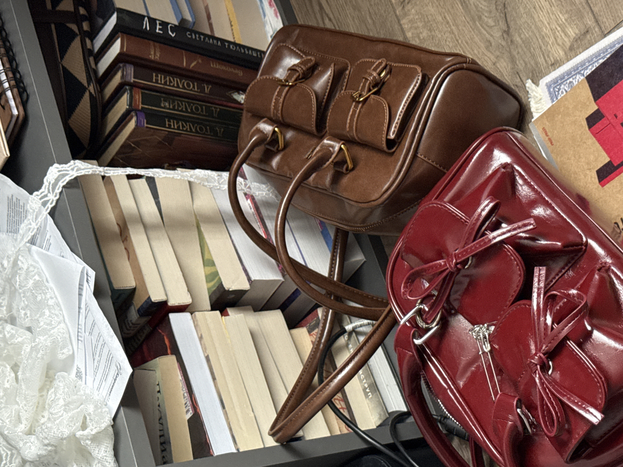 The image presents an indoor still life scene, possibly within a home or study, showcasing an arrangement of various objects on shelves and a wooden surface.
The primary subjects are two handbags and numerous books. In the upper left, a stack of books with dark covers is visible. The topmost book displays Cyrillic text: "ЛЕС" (LES, meaning "Forest") and "СВЕТЛАНА ТОЛ" (Svetlana Tol...). Beneath this, several books with dark spines show repeated inverted Cyrillic text, appearing as "НИВОЛ V" (NIVOL V). Adjacent to these stacked books, a spiral-bound notebook or diary with a dark, possibly wooden, cover is partially visible.
The central focus includes a structured brown handbag made of smooth, mid-brown material, likely leather or faux leather. It features two top handles and two prominent front pockets with buckled flaps, secured by gold-colored hardware. To the lower right, a glossy, deep red handbag is positioned. This bag is crafted from a shiny, patent-like material and adorned with decorative bows made from the same material. It also has a heart-shaped metallic zipper pull. A black cable is visible near this red bag.
Beneath the handbags and filling the lower half of the image are multiple shelves stocked with books, mostly displaying light-colored or beige spines, some white or darker, arranged vertically. A piece of white lace fabric lies in the lower left, partially covering some papers with illegible Cyrillic print, though "Капромекс" (Kapromeks) might be faintly discernible. A section of wooden surface is visible in the upper right corner, where a cardboard item with a red and black graphic design and faint "PROZ PRESS" text is located, along with a piece of blue patterned fabric.
The scene is illuminated by indoor lighting, and no people or specific activities are depicted. The visible Cyrillic text on the books aligns with the provided location context of Krasnodar, Russia.