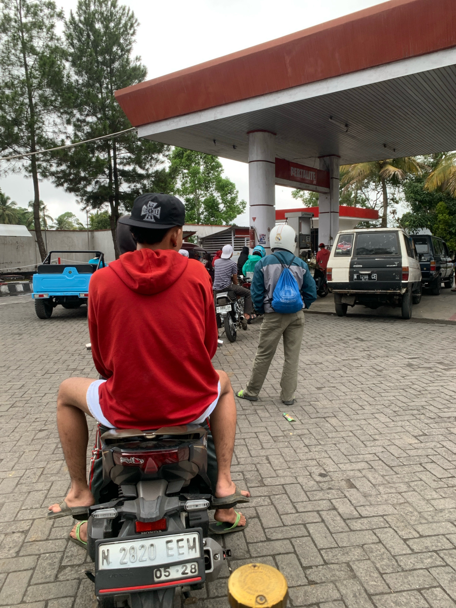In Pronojiwo, Indonesia, at a gas station, a person wearing a red hooded jacket and white shorts is seated on a motorcycle, viewed from behind. The motorcycle has a license plate reading "N 2820 EEM" and "05 28". To the right, another individual in a light blue and gray jacket, khaki pants, and a white helmet is standing, carrying a blue backpack. Behind them, a white and black two-tone SUV with license plate "N 1298 CE" and a black SUV are parked. A bright blue utility vehicle is parked to the left of the scene. The gas station canopy, with a red roof and gray ceiling, is visible overhead, and "PERTALITE" is displayed on a sign. The ground is paved with gray interlocking bricks. Tall trees are in the background.