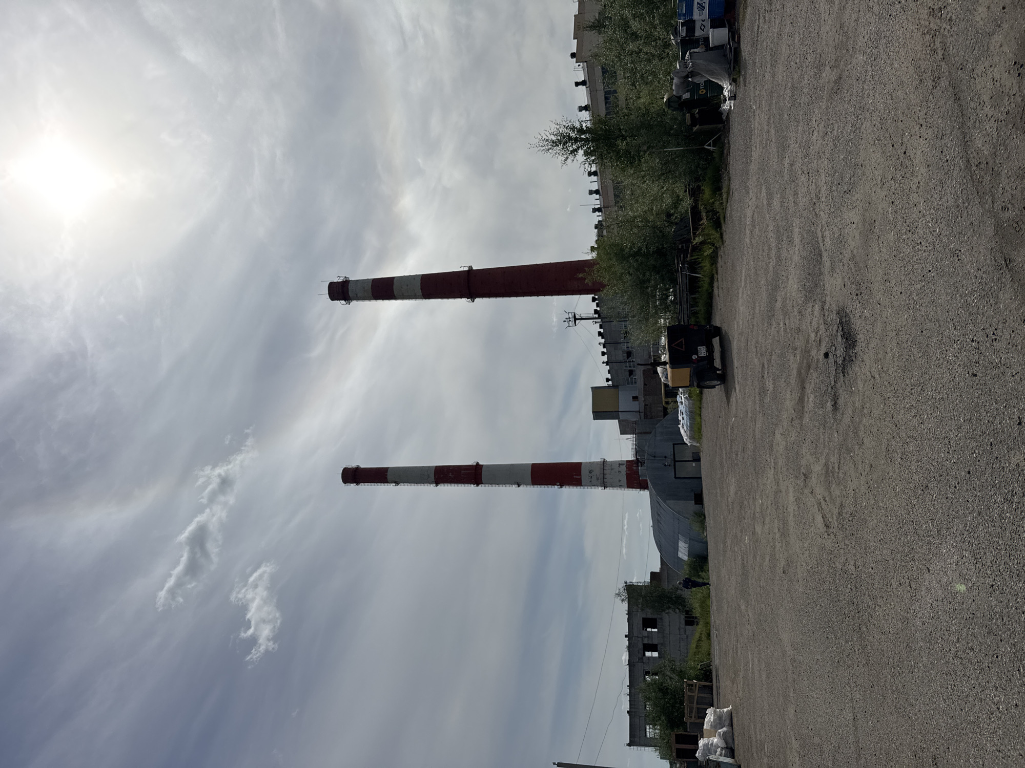The image depicts an industrial area, likely a power plant, in Usinsk, Russia, under a partially cloudy sky with bright sunlight. Two prominent smokestacks, painted with alternating red and white bands, dominate the background. The left smokestack is taller and appears to be slightly tilted. In the middle ground, a long, low industrial building with a curved metal roof is visible, along with other rectangular structures. Several vehicles, including a yellow trailer with a black canopy and a small utility vehicle, are parked in the foreground. Piles of white bags and various industrial materials are scattered throughout the scene, suggesting ongoing work or storage. A single person, dressed in dark clothing, walks in the distance near the curved-roof building. The ground is covered with gravel and asphalt, indicating a functional, if somewhat unkempt, industrial site. Lush green trees and bushes are interspersed around the buildings, providing a contrast to the industrial elements.