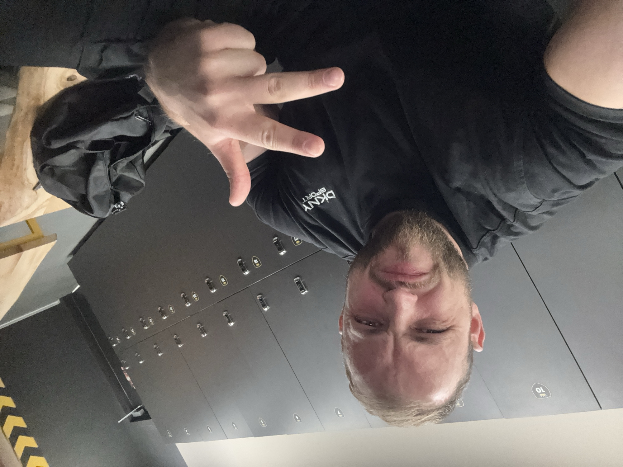 The rotated image captures a man taking a selfie in what appears to be a modern locker room or gym changing facility. The man, with a light beard and short, light-colored hair, is in the foreground, looking directly at the camera with a slightly squinted, determined expression. He is wearing a black short-sleeved t-shirt that displays the white "DKNY SPORT" logo on the chest. His right hand is raised, making a gesture with his index and middle fingers extended, resembling a V-sign.
In the background, a row of dark grey or black lockers with metallic handles extends along the wall. Several lockers have numbers and small, possibly leaf-like or wing-like, icons printed on them; "10" is clearly visible, with "09", "08", and "07" also discernible on adjacent lockers. To the man's left, a black bag or backpack is placed on a light wooden bench or surface. A section of floor with black and yellow diagonal hazard stripes is visible in the bottom left corner of the frame, further suggesting a facility with safety markings.