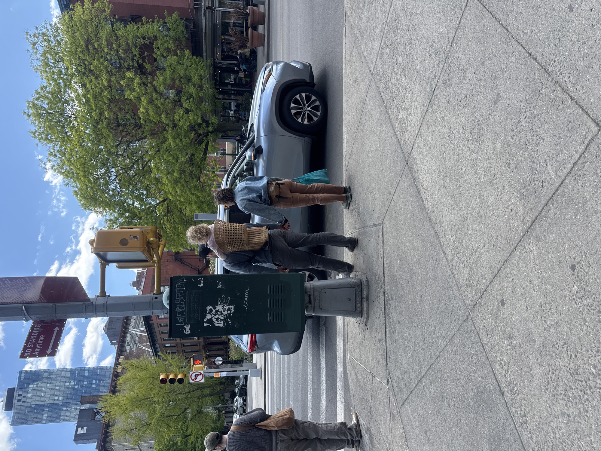 The photograph depicts a street scene in New York City, USA. The image is rotated counter-clockwise; the orientation should be adjusted.
In the foreground, there is a section of a sidewalk paved with rectangular slabs of concrete. In the middle ground, a traffic signal pole and a street sign are visible. A dark green metal control box attached to the pole is covered in graffiti. Further in the middle ground, there are three people waiting at a crosswalk. One person, facing away from the camera, is wearing a backpack made of wicker. Another person, partially obscured by a car, appears to be inspecting the vehicle. The third person is standing on the sidewalk, facing the opposite direction, carrying a shoulder bag.
A light-colored SUV is parked adjacent to the curb, partly obstructing the view of the crosswalk. In the background, various buildings and trees are visible, along with traffic lights and street signs. A "No Standing Anytime" sign is attached to the traffic pole. The sky is light blue with some clouds. The presence of brick buildings and foliage suggests a residential or mixed-use neighborhood.