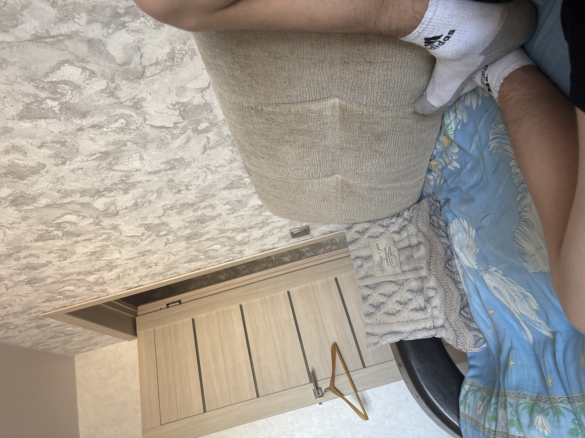 A first-person perspective shot shows a person lying on a bed with a blue patterned comforter. A beige couch is to the right, and a door with horizontal wooden panels is to the left. There is a wooden hanger hanging from the door handle. The walls are covered in textured wallpaper. The person is wearing white Adidas socks.