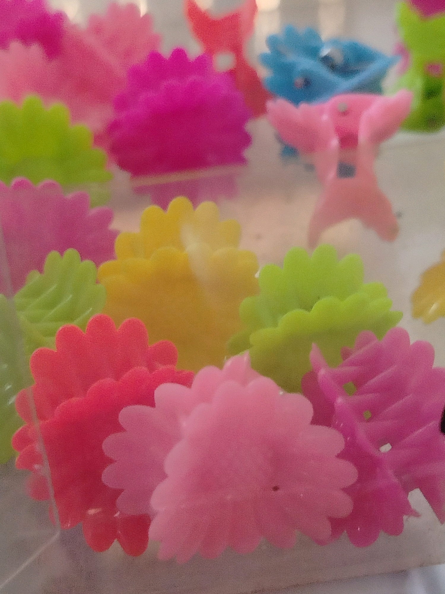 The image shows a close-up, slightly out-of-focus view of a collection of colorful plastic hair clips. The clips are primarily in shades of pink, yellow, and green, with some blue clips visible in the background. They appear to be in the shape of small flowers or decorative pieces with scalloped edges. The lighting is bright, and the background is blurred, suggesting the clips are likely displayed in a store or a personal collection. There are no people or animals visible in the image. The scene does not provide any specific information or visual cues that would associate it with the city of Көлкент, Kazakhstan.