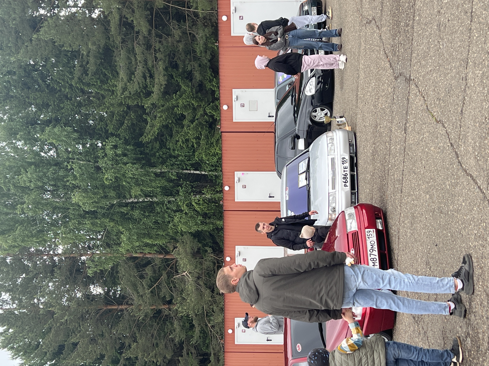 The image depicts an outdoor car gathering in the city of Прикамский, Russia, featuring several individuals and lowered vehicles on an asphalt lot. In the foreground, a tall adult male in a dark olive green hooded jacket and light blue jeans stands, holding the hand of a young child wearing a green vest and dark beanie. A red car, possibly a Lada, with Russian license plate "M879MO 159" is partially visible in front of them, appearing lowered. Behind them, a silver car, also resembling a lowered Lada, displays Russian license plate "P686TE 159" and has a golden trophy placed on the ground nearby. Further right, a black lowered car, likely a Lada, with multi-spoke wheels is parked. Several individuals are standing around these vehicles; this includes an adult male in a black jacket holding a light-colored beanie, a young woman in a pink hoodie, and another young woman in a dark jacket and brown vest. Two additional males are visible in the mid-background near a dark green car with a license plate ending "OK 159". In the background, a line of five identical reddish-orange modular buildings with white doors numbered 10 through 14 are visible. Behind these structures, a dense forest of various trees forms the ultimate backdrop under an overcast sky. The ground is cracked asphalt pavement.