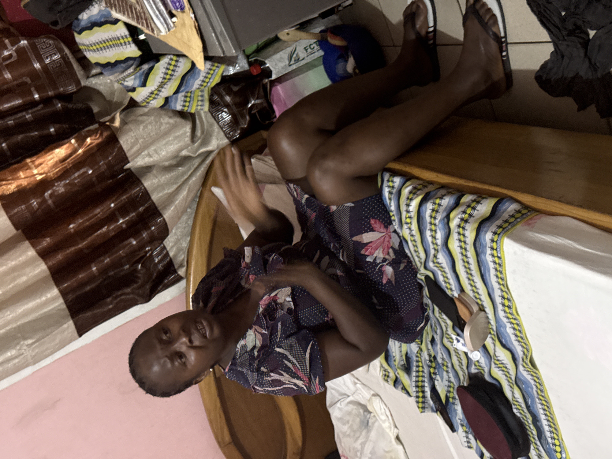 An adult Black woman is sitting on a bed with her legs extended towards the upper-right corner of the image. She wears a patterned dress and flip-flops. A patterned bedsheet is partially visible. Various items including boxes, fabric, and clothes are scattered around the bed. The wall is pink. The location is potentially a residential space in Abuja, Nigeria.