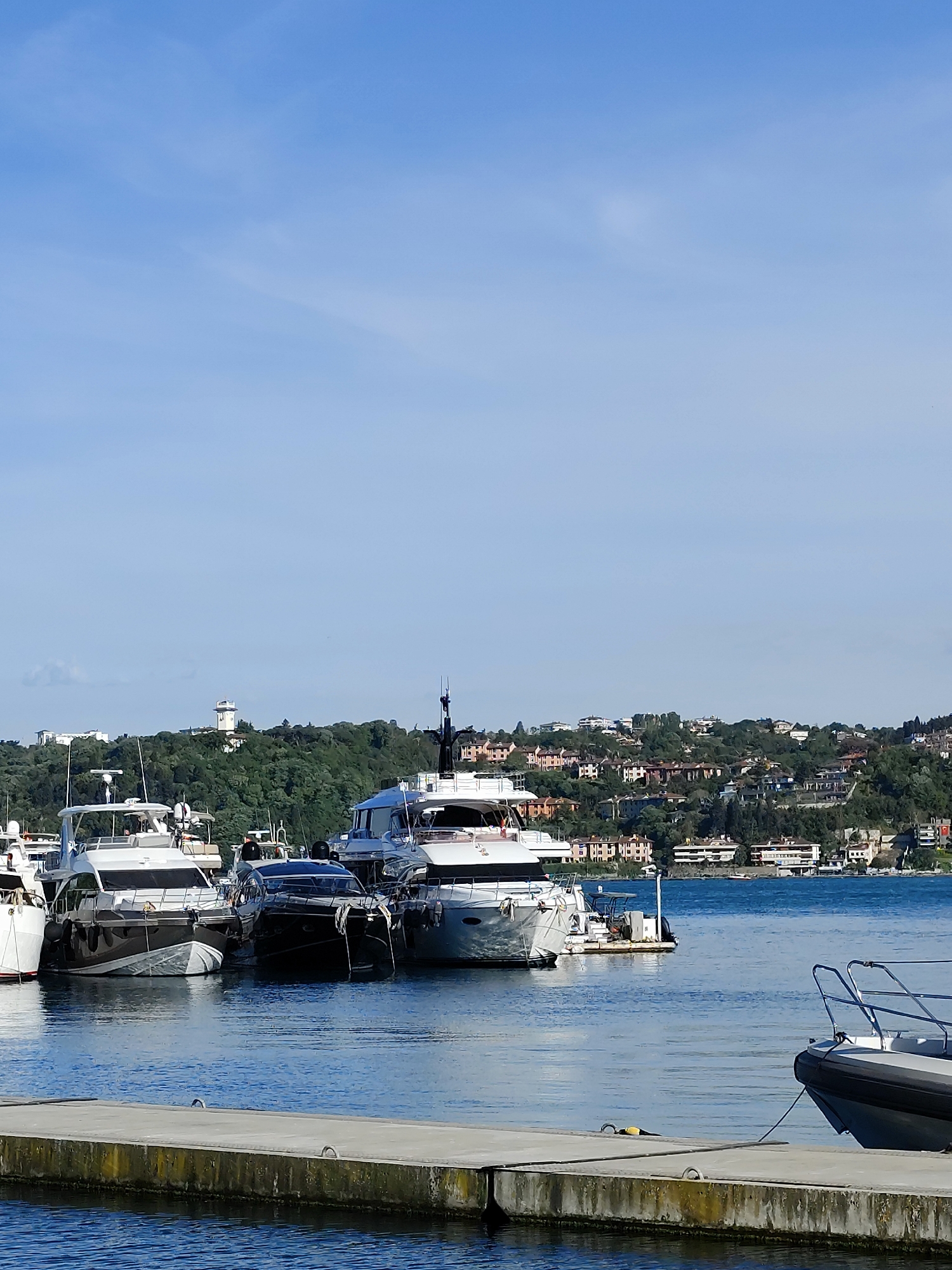 The image shows a marina on the Bosphorus, located in Sarıyer, Turkey. Several luxury yachts are docked alongside a concrete pier. The water is calm and blue. In the background, a green hillside is dotted with houses and a white tower-like structure. The sky is blue with some light clouds. Part of a small boat appears in the foreground on the right.