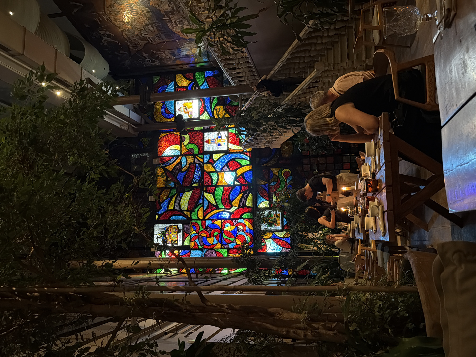 This image depicts the interior of a restaurant in Moscow, Russia. The scene is illuminated by ambient lighting and the vibrant colors of a large stained-glass window featuring abstract geometric shapes and what appear to be miniature cityscapes.
In the foreground, a wooden table is set with place settings, including cups, saucers, a teapot, and a lit candle. A woman with blonde hair is seated at this table, leaning on her hand, facing away from the camera and towards the stained-glass window. A man with grey hair is seated next to her, also facing away.
Further back, another table is occupied by a couple. The man, wearing a dark t-shirt and apron, is serving or interacting with the woman, who is wearing a black top. They appear engaged in conversation.
The restaurant environment is lush with indoor plants, including large trees and potted greenery, creating a semi-tropical atmosphere. To the right, a grand, ornate staircase with carved balustrades ascends, with a person visible on the stairs. A mural or tapestry is visible on the wall to the right of the staircase. The overall impression is of a sophisticated and atmospheric dining space.