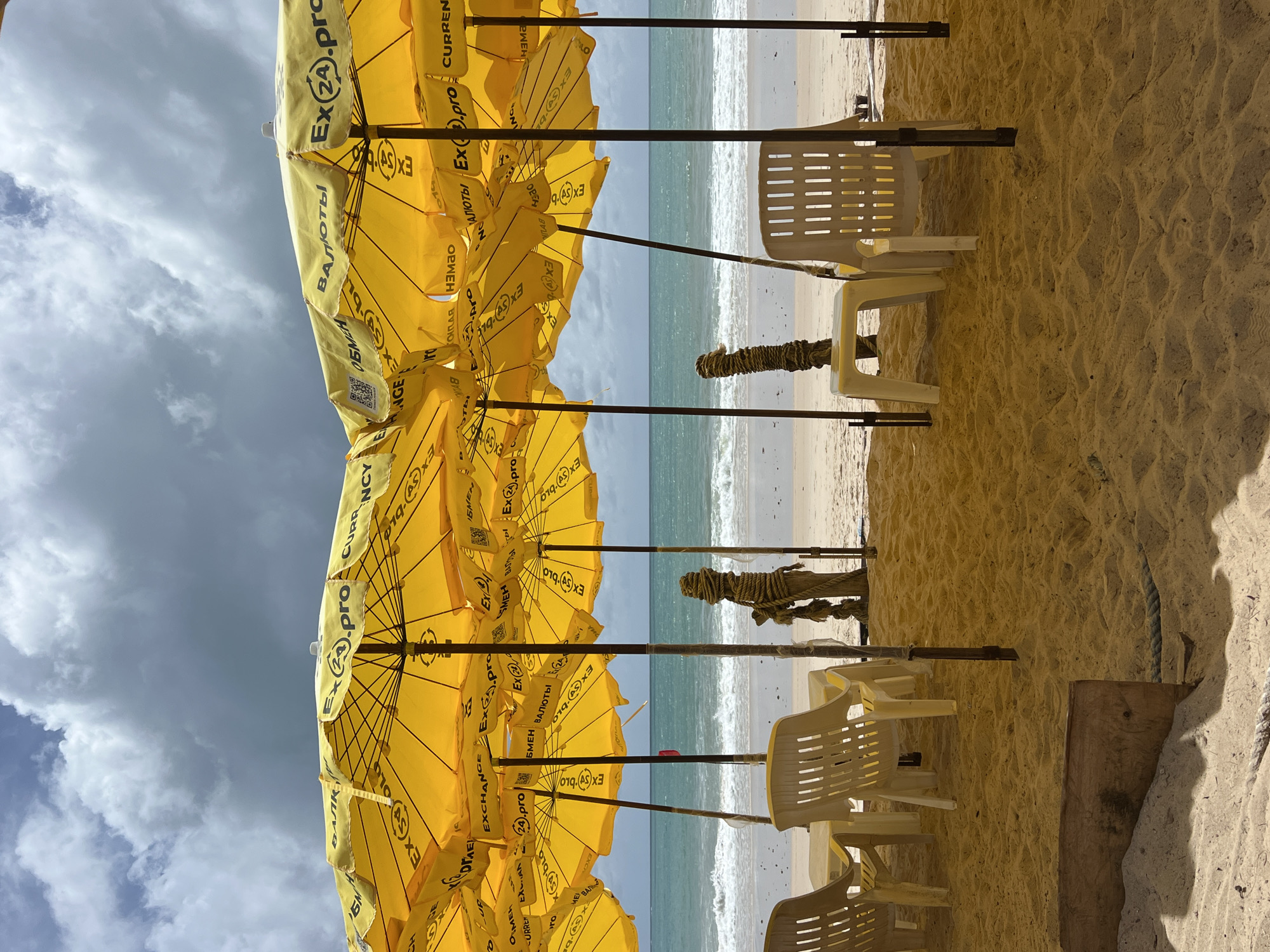 The image shows a beach scene with rows of yellow beach umbrellas and white plastic chairs. The umbrellas are printed with "Ex(24).pro" and various currency exchange-related words in both English and what appears to be Russian. The ocean is visible in the background, with small waves breaking on the shore. The sky is partly cloudy. The sand is beige and textured.