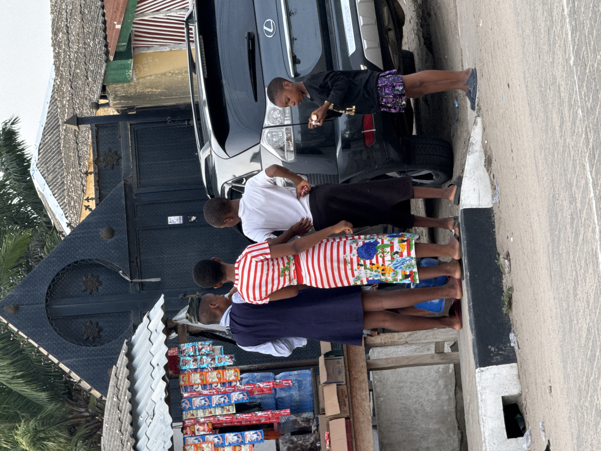 The image depicts an outdoor scene in Badagry, Nigeria. Four young individuals are near a black Lexus SUV parked on the side of a paved road. One individual is at the rear of the vehicle, near the taillights, and seems to be holding a small object. Three other individuals stand near a stall. This stall displays stacks of small product packages. A grey corrugated metal roof covers part of the stall, and a dark metal gate is adjacent to it. The surrounding environment includes trees and other buildings in the background.