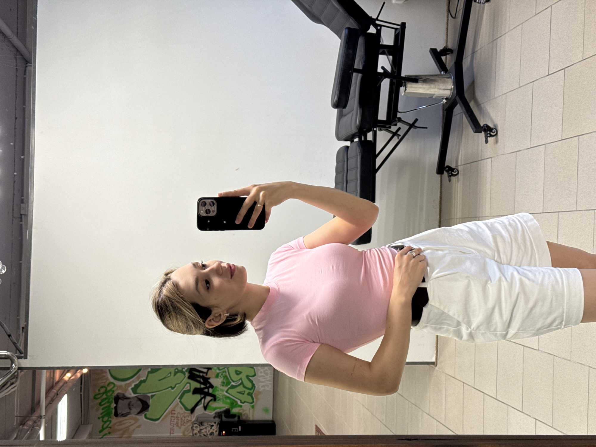 A young woman with blonde and dark brown hair, wearing a pink t-shirt and white shorts, is taking a mirror selfie in what appears to be a tattoo studio. She is holding a black smartphone in her right hand, capturing her reflection. The background shows a white wall with graffiti art on the left, and a black tattoo chair with armrests and wheels on the right. The floor is tiled in a light beige color. The lighting is bright, suggesting an indoor setting.