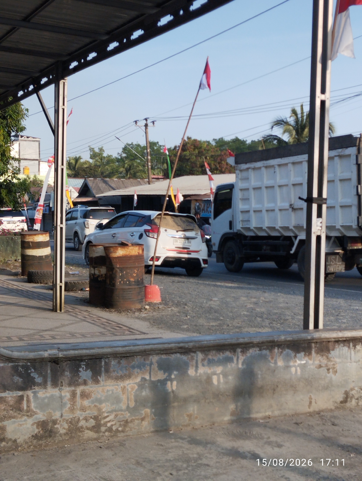 The image captures a street scene in Serongga, Indonesia, under a clear blue sky. In the foreground, a worn concrete wall shows signs of peeling paint and weathering. Two large, rusty metal barrels are positioned on the side of a paved road. One barrel is on the left, and the other is to its right, with a red cone-shaped object near it. A tall pole with black and white stripes stands between the barrels, supporting wires that stretch across the scene.
Further down the road, several vehicles are visible. A white dump truck is parked on the right, its white cargo bed featuring a grid pattern. To the left of the truck, a white SUV is parked, and behind it, another white SUV is also present. A smaller white car is seen further in the distance on the left.
Several flags are displayed throughout the scene. Red and white flags, likely the Indonesian flag, are mounted on poles. One prominent flag is positioned behind the metal barrels. Smaller flags are also visible on poles in the background. Trees and buildings with corrugated roofs are scattered throughout the midground. The overall impression is of a roadside setting, possibly an area with some commercial activity or residential buildings. A timestamp in the bottom right corner reads "15/08/2026 17:11".
