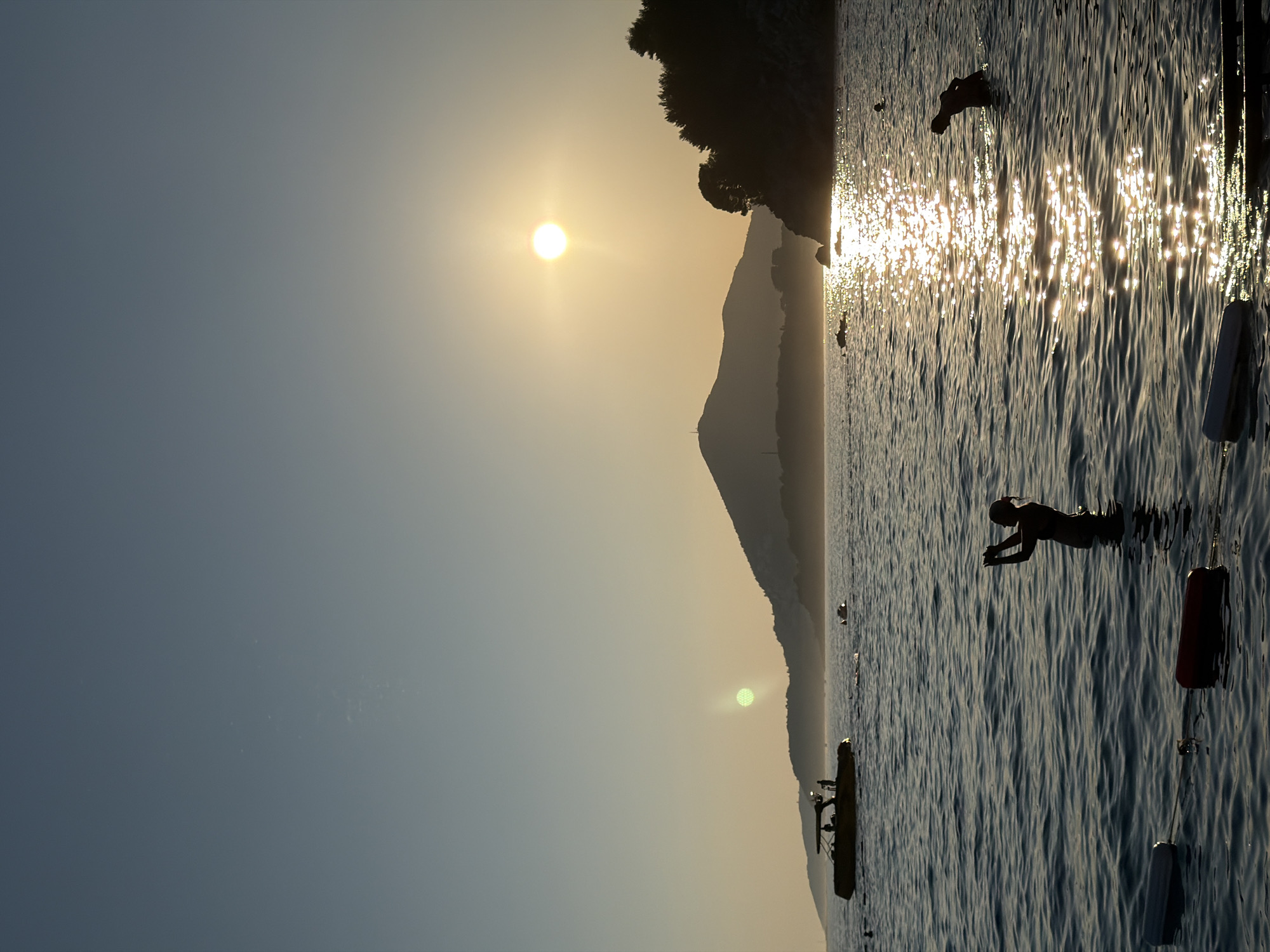 The image depicts a serene sunset scene at a beach in Boreti, Montenegro. The sun, a bright orb, is low on the horizon, casting a golden sheen on the gently rippling water. Silhouettes of people are visible enjoying the sea. In the foreground, a person stands in the water, seemingly taking a photograph with their phone. Further out, another person is wading in the water. A boat with several individuals on board is positioned to the left of the frame, near the shore. The background is dominated by a mountainous silhouette, characteristic of the Montenegrin coastline, with lush green trees adorning the slopes. The sky transitions from a clear, pale blue at the top to a warm, orange hue near the horizon, indicative of the setting sun. Floating buoys are strung across the water in the foreground, marking a designated swimming area. The overall atmosphere is tranquil and idyllic, capturing a moment of natural beauty and human activity at dusk.