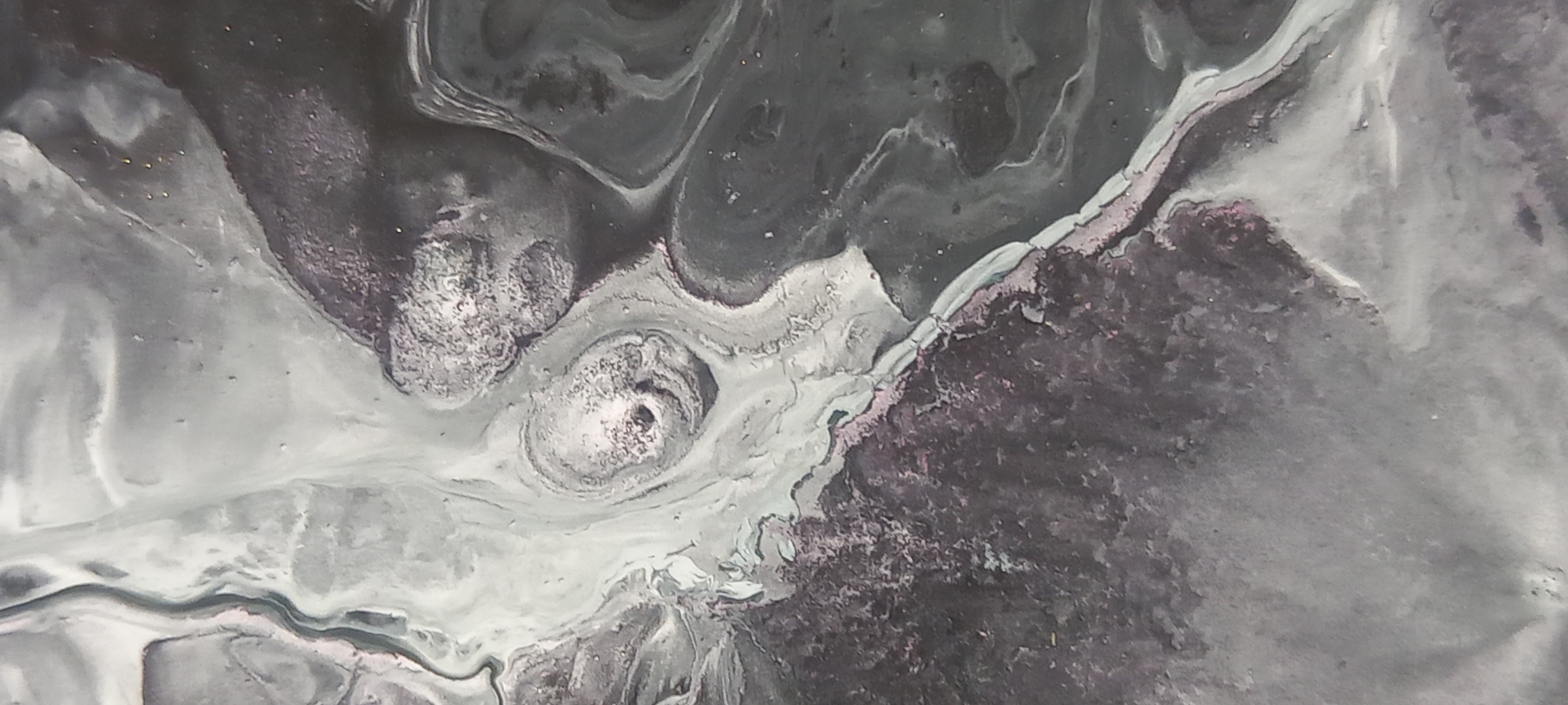This image is an abstract close-up of a fluid art painting. It features swirling patterns of grey, white, and black, with some subtle pink and glitter accents. The textures are varied, ranging from smooth, flowing lines to rough, granular areas. There are no identifiable landmarks or features that would suggest a specific location within Kryvyi Rih, Ukraine. The image is purely an artistic representation of abstract forms and colors.