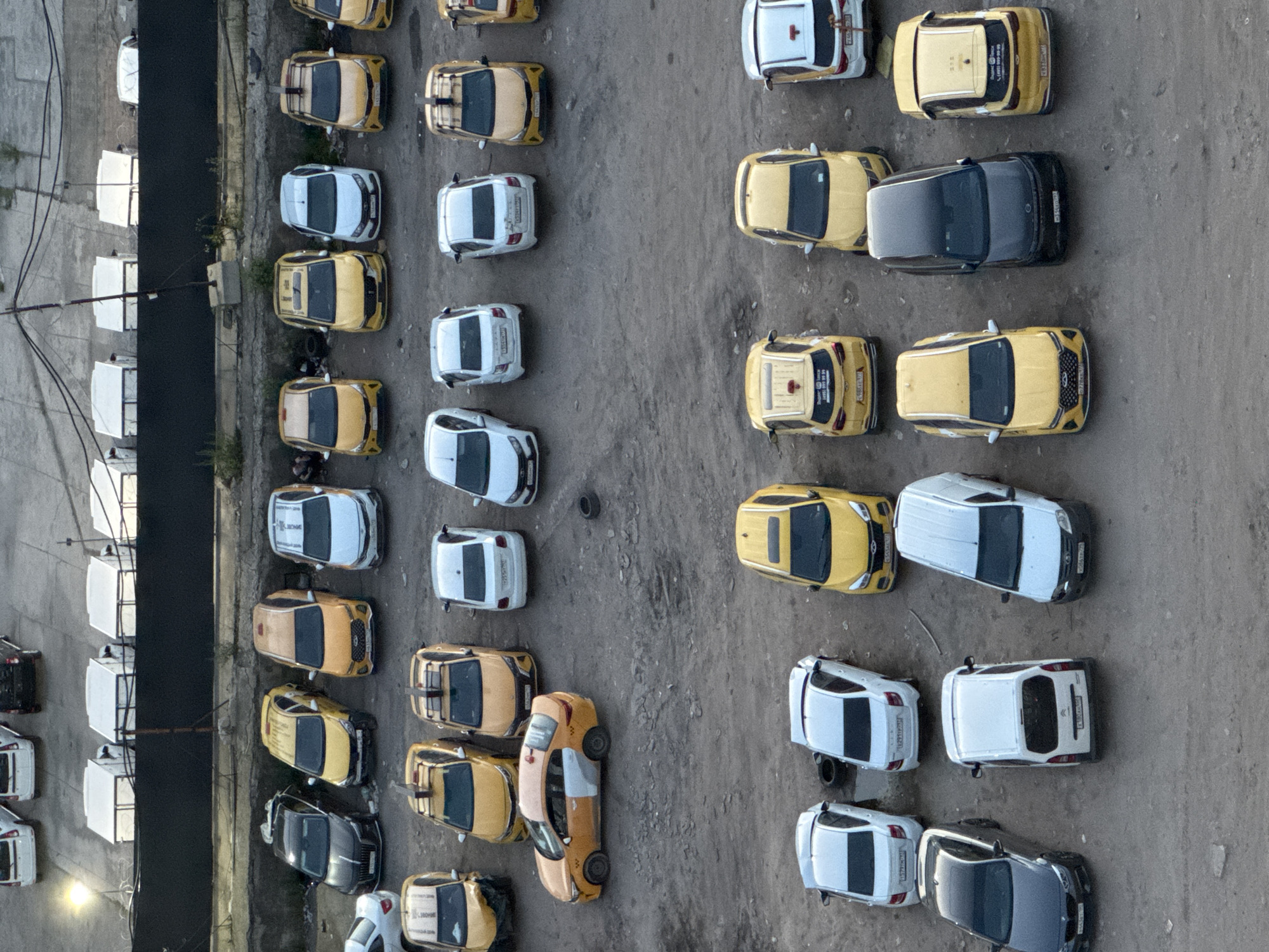 The image is an aerial view of a parking lot filled with numerous yellow and white cars, many of which appear to be taxis. The cars are parked in neat rows on a gravel surface. In the background, there are several white modular structures lined up against a dark fence. A few trees and bushes are visible near the fence. The overall impression is of an organized fleet of vehicles, possibly awaiting dispatch or maintenance. The scene suggests a functional, utilitarian space rather than a residential or commercial area.