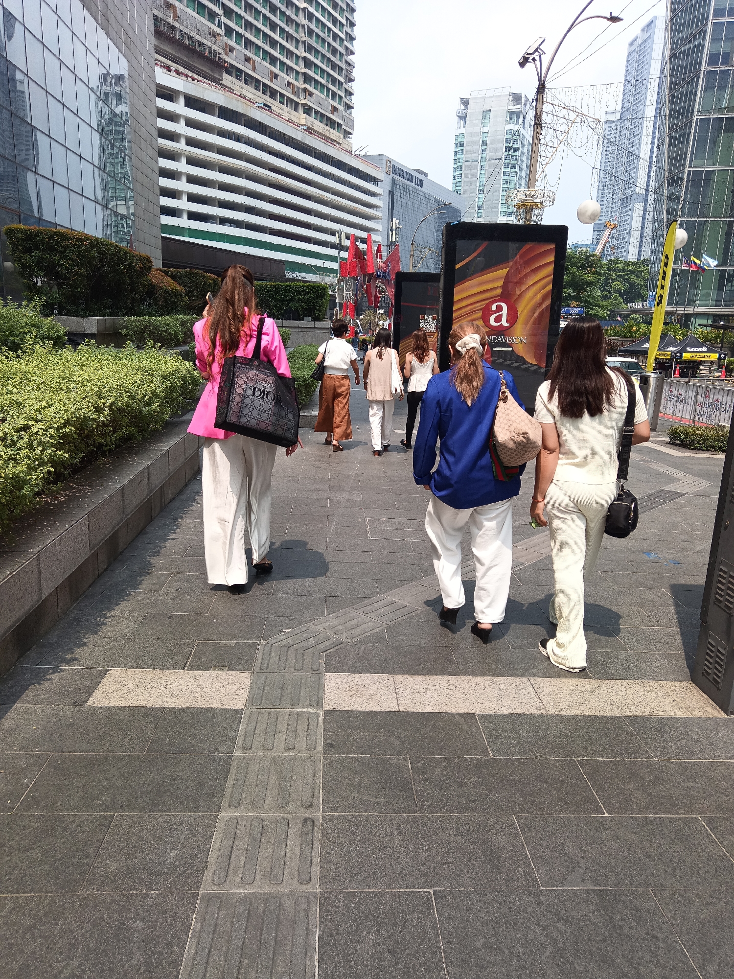 The image depicts a street scene in Kuala Lumpur, Malaysia, likely within the city center or a commercial district. In the foreground, a woman in a bright pink blazer and wide-leg white pants, carrying a black Dior tote bag, walks away from the camera. She is followed by three other individuals. The woman to her right wears a blue jacket and white pants, carrying a beige and green patterned bag. The woman to her right wears a cream-colored textured set with a black crossbody bag. Behind them, a fourth person in white pants and a cream top is also walking away. The environment consists of a paved walkway with tactile paving for the visually impaired. In the background, modern skyscrapers with glass facades dominate the skyline. One building appears to be under construction. A large digital billboard advertising "UNDAVISION" is prominently displayed. The scene is characteristic of a busy urban environment in Kuala Lumpur.