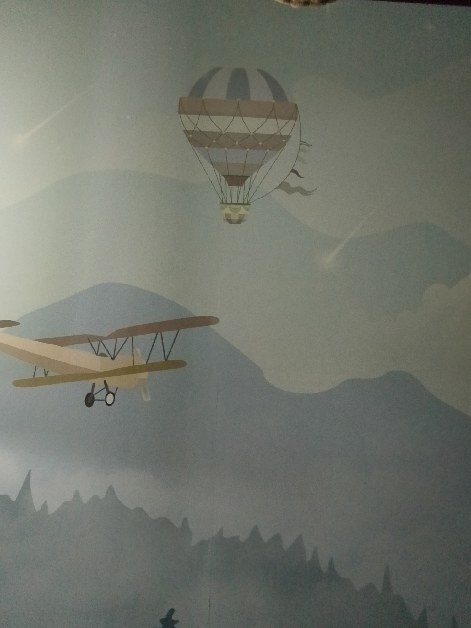 This media file depicts a mural or artwork featuring an airplane and a hot air balloon in a mountainous landscape. The primary subjects are the biplane in the foreground and the hot air balloon in the upper portion of the frame. The background consists of stylized, layered mountains, fading into a soft, blueish-gray sky. Light streaks, possibly representing shooting stars or atmospheric effects, are visible in the sky. The biplane is a light tan color with brown wings and black struts. It has two wheels and a propeller. The hot air balloon has a striped design, with segments of tan and light blue. It is suspended by ropes from a basket. The scene evokes a sense of adventure and flight, with an implied time of day that could be dawn or dusk due to the soft lighting and muted colors. The location cues suggest a fantastical or artistic representation rather than a realistic depiction of a specific place in Kalachinsk, Russia. No text is visible in the media file.