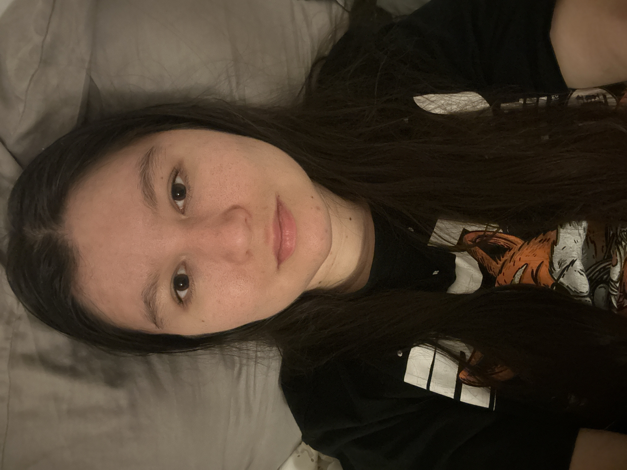 A person with long, dark hair is lying down, facing the camera. The person has fair skin and brown eyes. The person is wearing a black t-shirt with a design on it. The person is lying on a grey pillow. The location is not visible in the image.