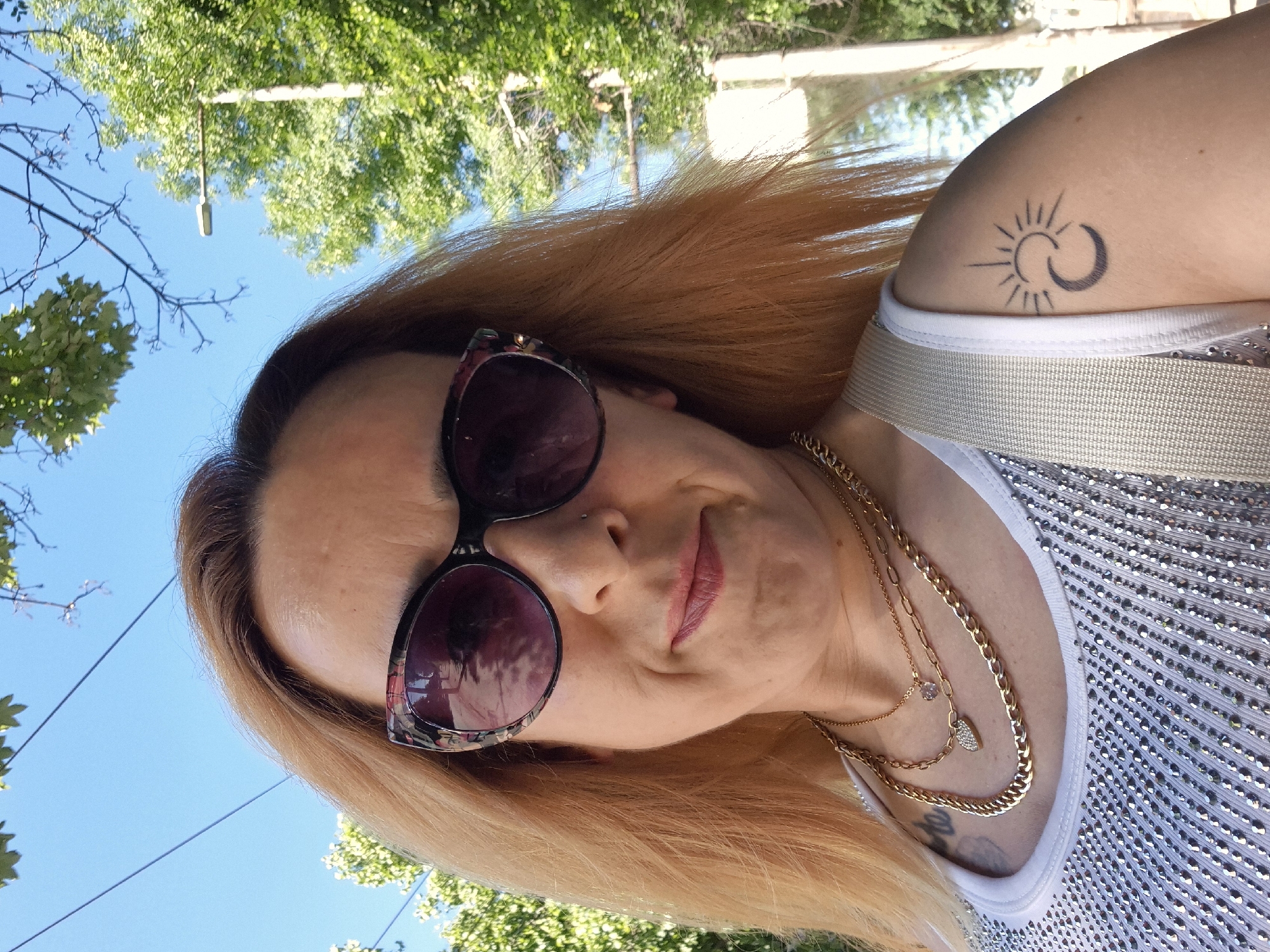 A woman with shoulder-length, auburn hair, wearing large, patterned sunglasses and a white tank top embellished with numerous small, reflective rhinestones, is pictured outdoors. She has a small nose ring and wears two gold necklaces, one with a heart-shaped pendant. A tattoo of a crescent moon and a sun is visible on her left shoulder. The background consists of a clear blue sky, green trees, and utility lines. The woman is looking towards the camera with a slight smile. The setting appears to be urban, possibly a street or park, given the presence of trees and utility poles.