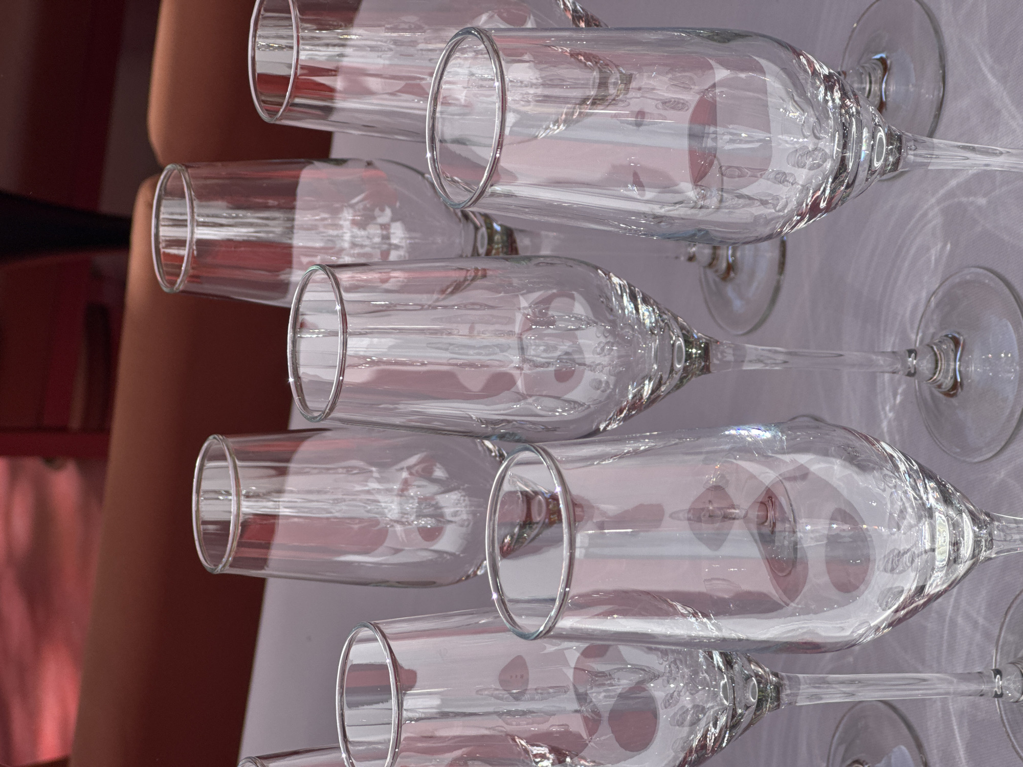 The image shows a close-up of several clear champagne flutes lying on their sides on a white tablecloth. Light creates patterns on the surface, reflecting through the glass. In the background, out of focus, is a red surface. There is no information to indicate the location in Yerevan, Armenia.