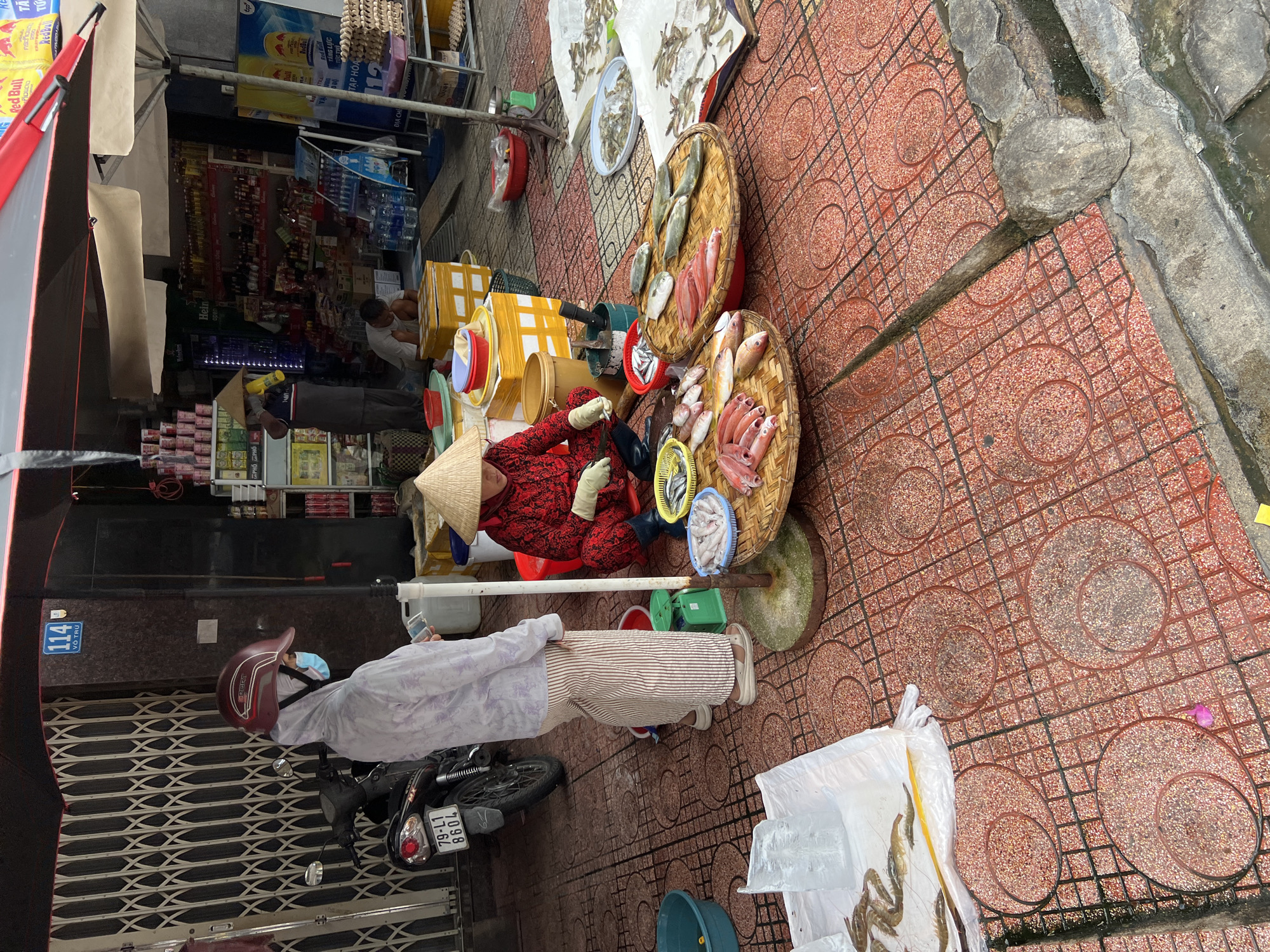 A street market scene in Nha Trang, Vietnam, features a vendor in a traditional conical hat selling fresh fish. The vendor, wearing a red patterned top and black pants, is seated behind a display of fish arranged on woven mats. Several types of fish, including some appearing to be snapper and smaller silvery fish, are visible. To the left, a person wearing a hoodie, a helmet, and a face mask stands near a motorcycle with a license plate reading "79-L1 8604". The market is set up on a red tiled sidewalk with a drainage channel running along the side. In the background, a store selling various goods is visible. An umbrella shades the market stall.