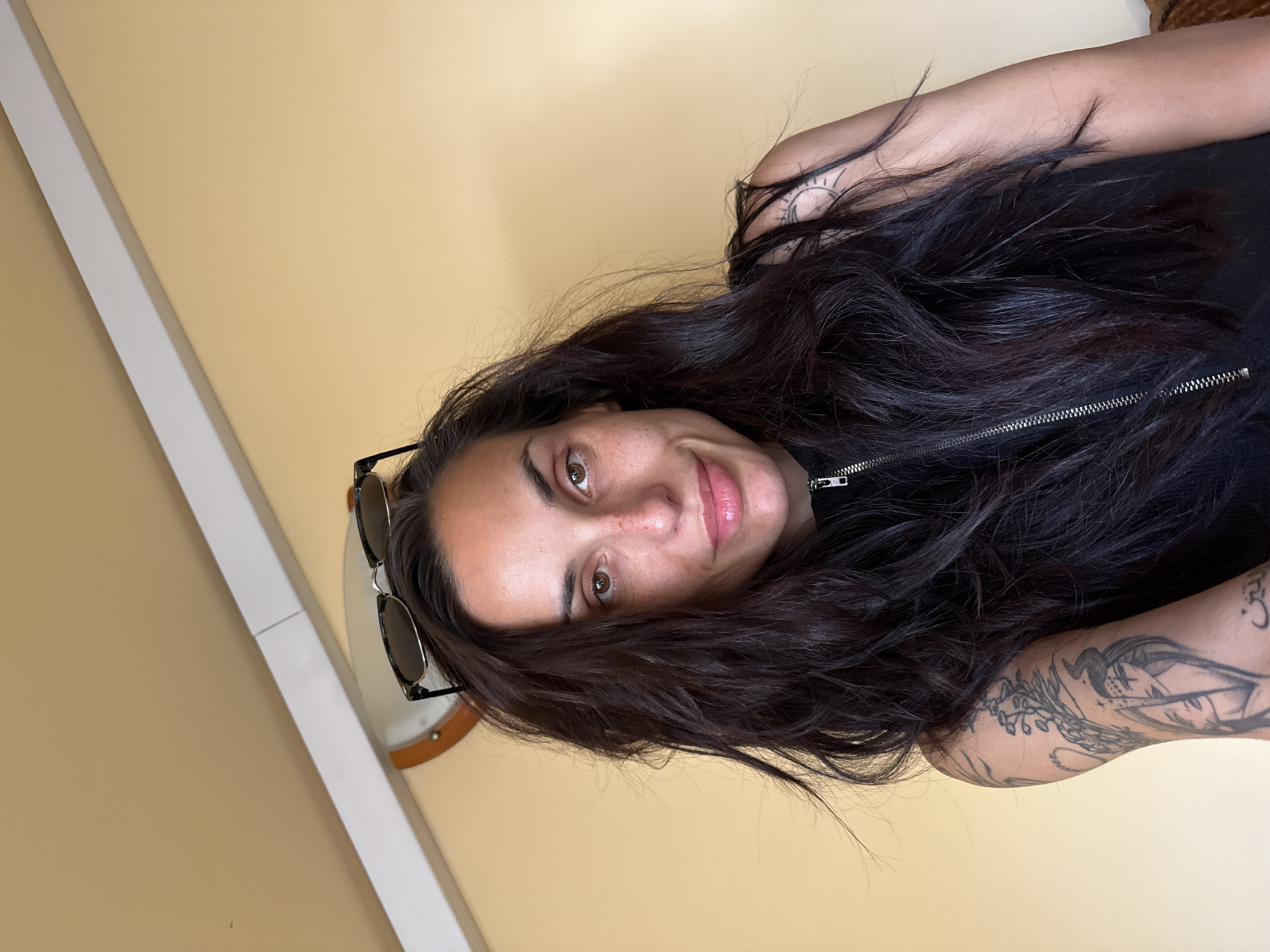 A medium shot of a person with long, dark hair partially obscuring their face, taken indoors with a beige wall as a background. The person has sunglasses perched on their head, above their forehead. They are wearing a black zippered top. Tattoos are visible on both arms. The image lacks contextual clues to determine the exact location in Pleven, Bulgaria.