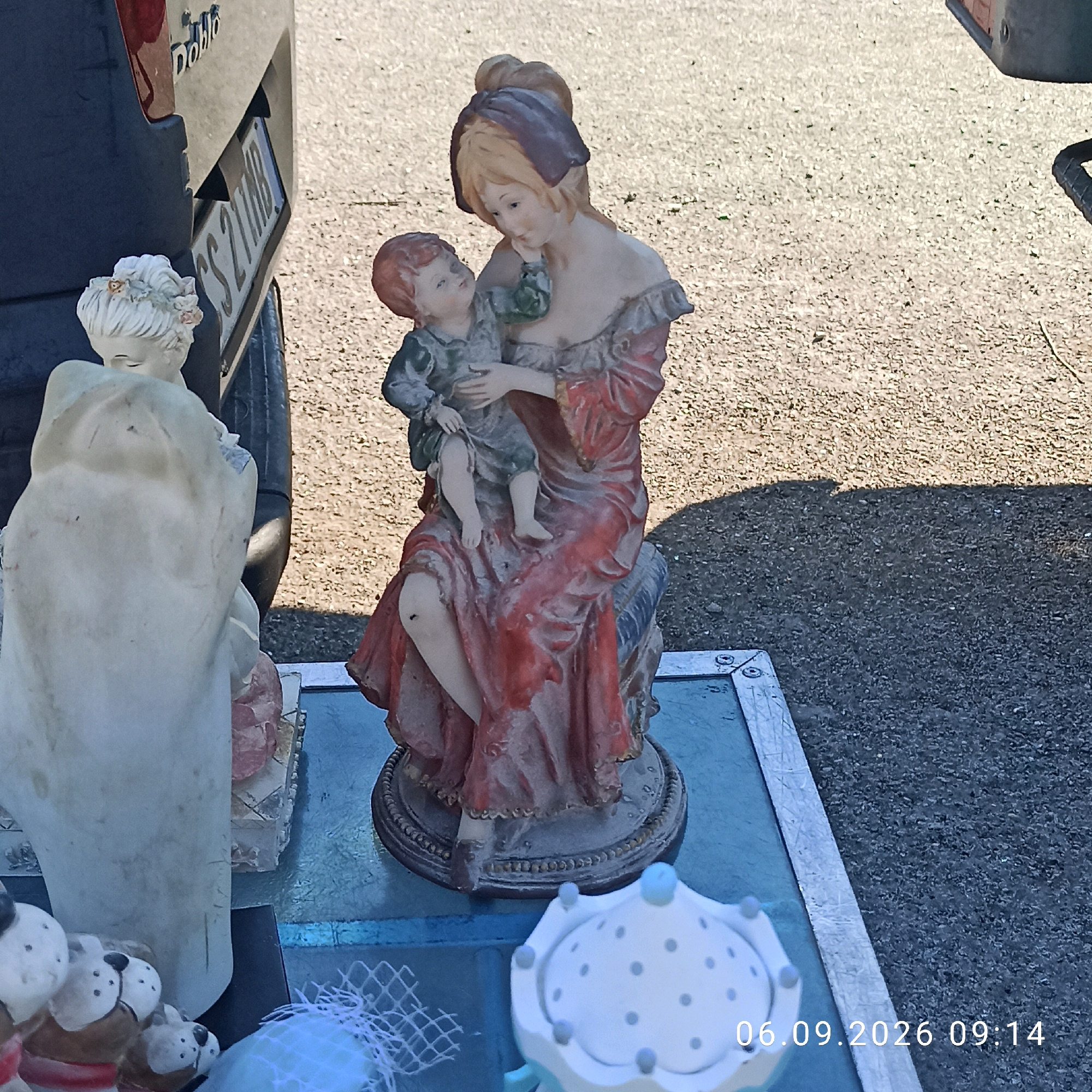A statue of a woman cradling a child is displayed outdoors, likely at a market or street stall in Sannicandro di Bari, Italy. The statue, with faded colors, depicts the woman in a red dress with ruffled sleeves and a purple headscarf, holding a child in a blue garment. To the left of the main statue is a partially visible, lighter-colored statue, possibly of an angel or another figure. In the foreground, to the right, is a small white decorative object with gray polka dots and light blue accents. The background consists of a gravelly surface with a shadow cast from an unseen object. A vehicle's bumper and license plate (bearing the characters "SS 211 MB") are visible to the left, indicating its proximity to a road or parking area. The date "06.09.2026 09:14" is superimposed on the lower right of the image.
