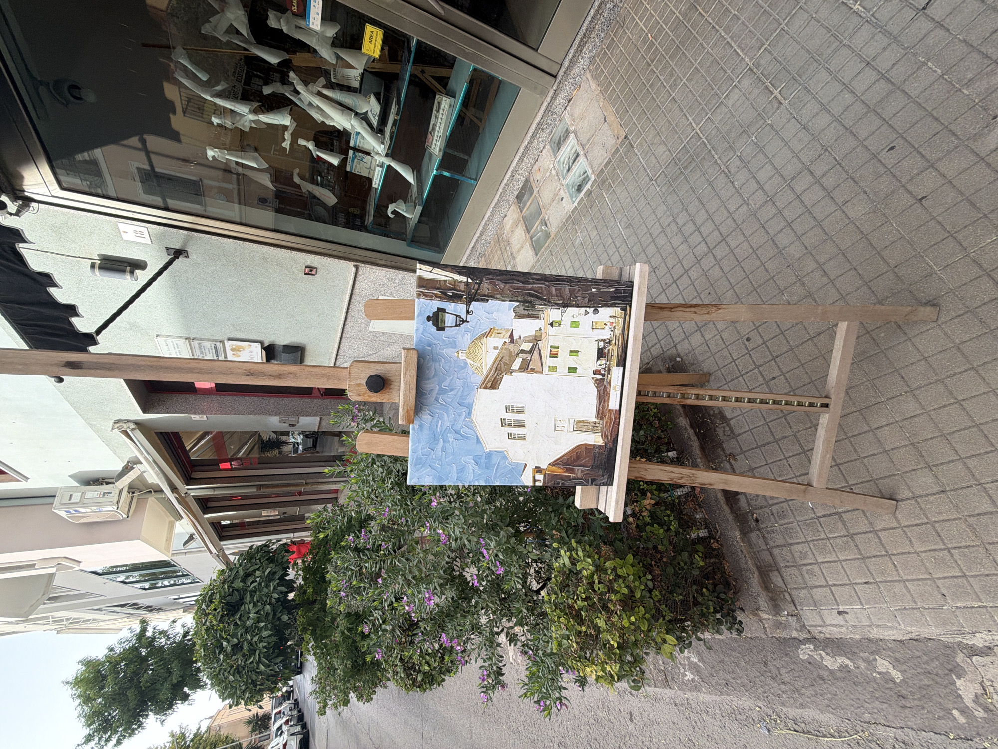 This media file displays an oil painting on an easel set up on a paved sidewalk. The painting depicts a street scene with white buildings, a blue sky, and a dome, possibly a church or historical landmark, illuminated by sunlight. A street lamp hangs over the scene. The painting is placed in front of a storefront with large windows displaying white mannequins. Greenery, including flowering bushes and trees, is visible in the foreground and to the left of the easel. The sidewalk is paved with square tiles and a portion of a street with parked cars is visible in the background. The lighting suggests it is daytime, possibly late morning or early afternoon, with clear weather. The scene evokes a sunny Mediterranean atmosphere, characteristic of a location like Alghero, Italy. No people are visible in the frame. The overall impression is one of an artist displaying their work outdoors, capturing the essence of the local architecture and ambiance.