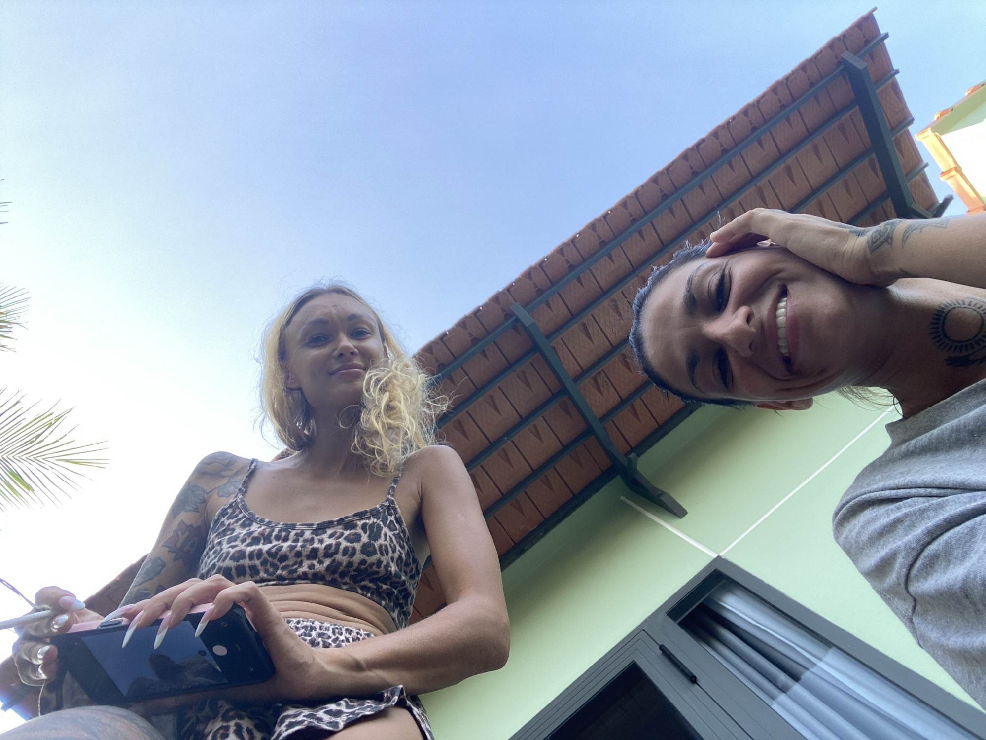 Two women are taking a selfie outdoors. One woman in the foreground is wearing a grey sweatshirt and has her hand on her face. She has dark hair and a tattoo on her chest. The second woman is in the background wearing a leopard-print top and has blonde hair. She is holding a smartphone. The background shows a light blue sky, a light green building, and a tiled roof. A palm frond is visible in the upper left corner of the image. The scene appears to be in a tropical or subtropical location, possibly Mũi Né, Vietnam, given the presence of palm trees and a clear sky.