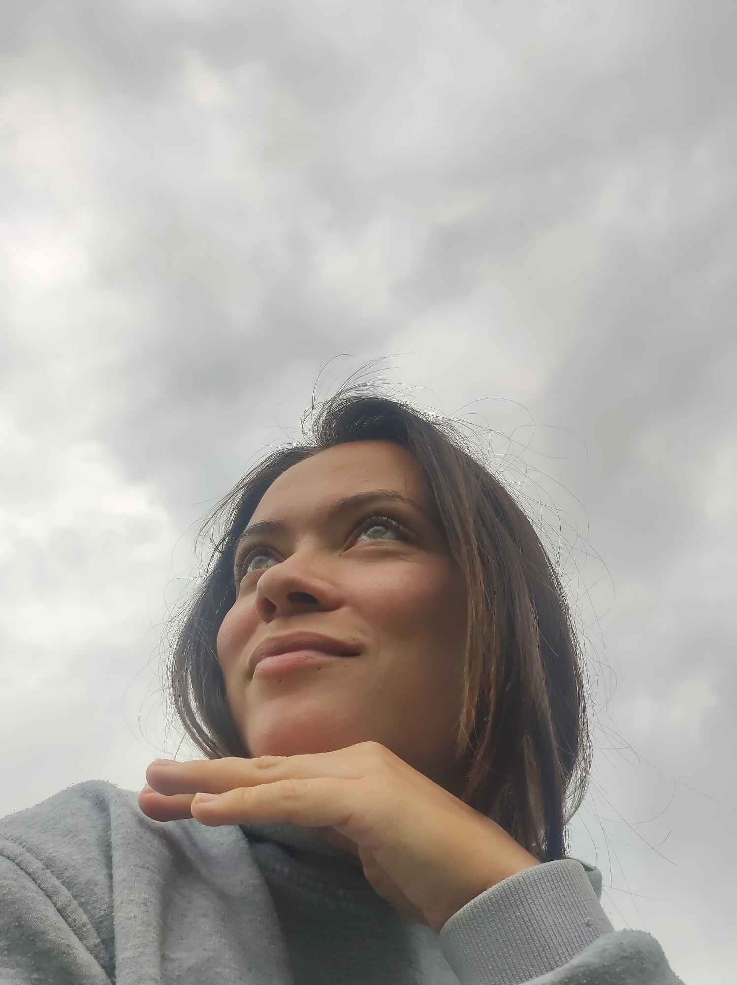 A close-up, low-angle portrait captures a young woman looking upwards against an entirely overcast sky. The woman, appearing to be in her 20s or 30s, has medium-length dark brown hair and light-colored eyes, which are directed towards the sky. A soft, serene smile is visible on her lips, and her right hand rests gently under her chin, suggesting a thoughtful or contemplative expression. She is wearing a grey hooded sweatshirt, visible on her shoulder and arm. The background is a dense, uniformly cloudy grey sky, indicating overcast weather during the daytime. No other objects, structures, or text are visible within the frame. The image conveys a sense of introspection or looking towards the future, set in Фоки, Russia.
