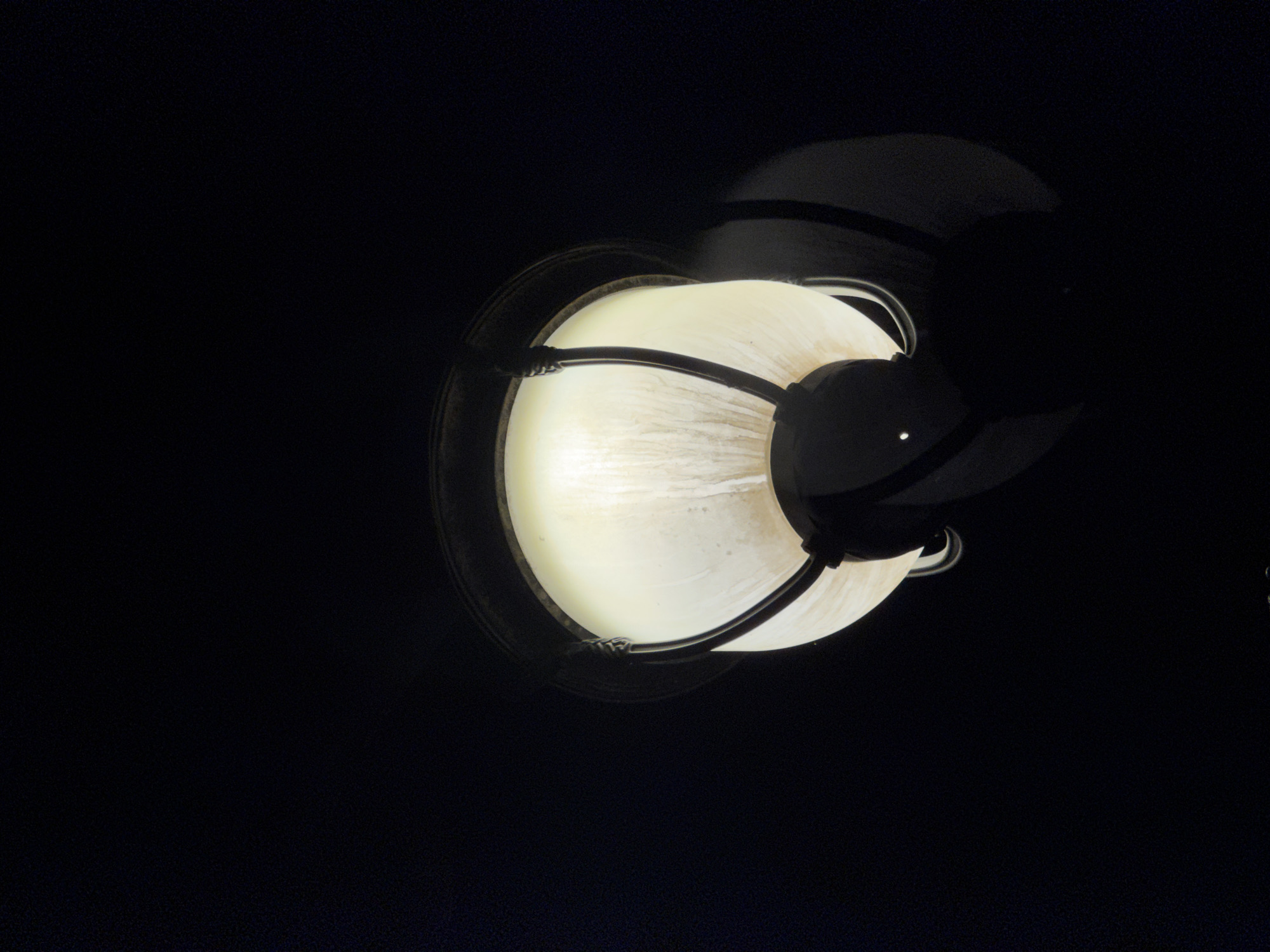 A low-angle, close-up shot captures a lit outdoor lantern against a dark background. The lantern has a frosted glass shade with visible vertical lines and is held by a dark metal frame with decorative, curved elements. The light source inside the lantern emits a warm, yellow glow. The overall impression is of a single, illuminated object in darkness, typical of street lighting in urban environments. This image does not provide any specific visual cues to determine its location within Tashkent, Uzbekistan.