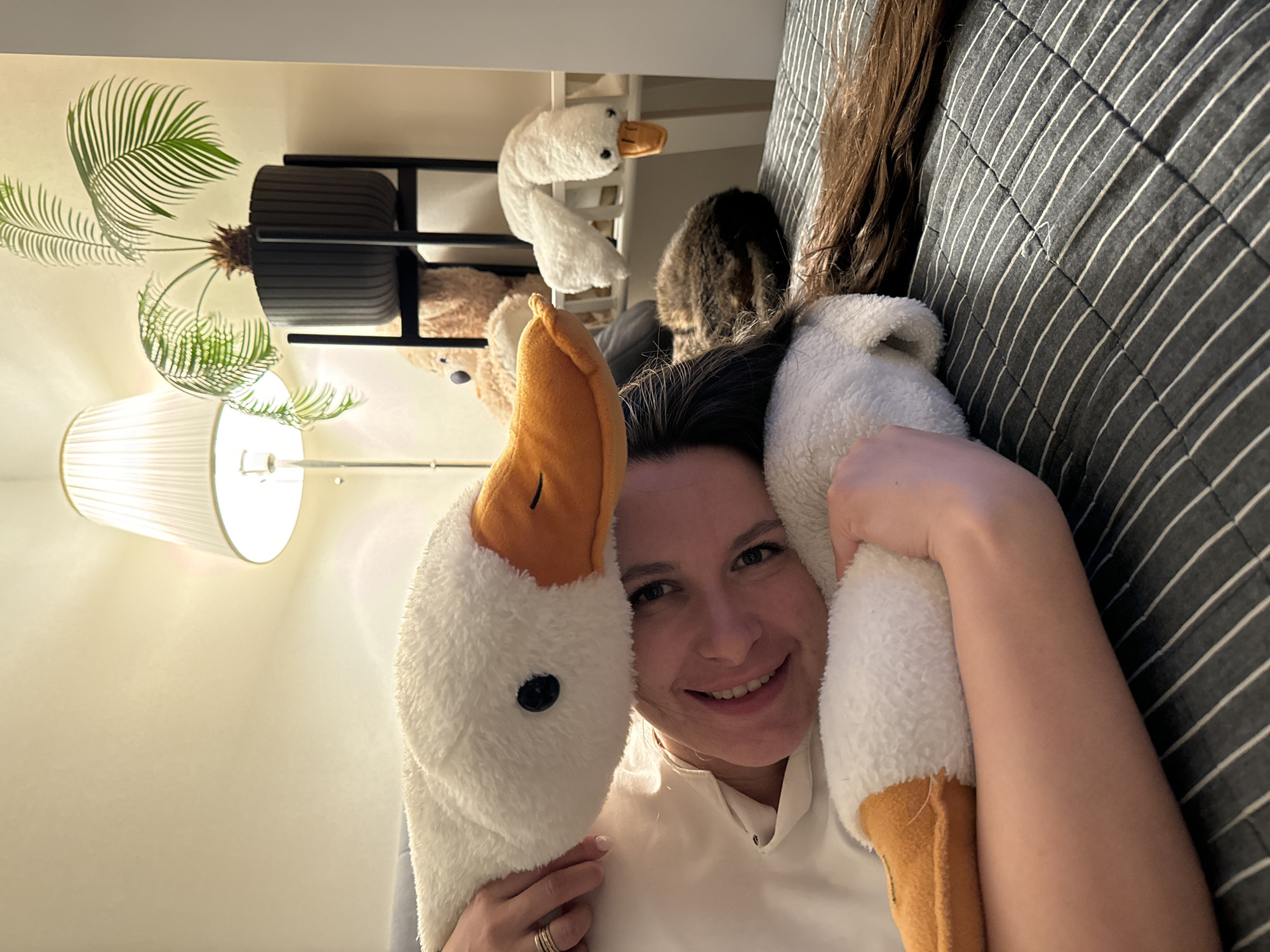 An indoor, portrait-oriented photograph depicts a smiling woman lying down, looking directly at the camera. She is playfully framed by two large, plush white goose heads, one on each side of her face, featuring orange beaks and black eyes. The woman wears a white top and a multi-banded ring on her left hand. Her dark brown hair cascades to the right.
To her right, a dark grey blanket with white stripes is visible. In the background to her left, an indoor plant, possibly a sago palm, sits in a dark ribbed planter on a black metal shelf. A floor lamp with a white pleated shade illuminates the scene from the left. Above the plant, another smaller white plush goose toy is perched. A fluffy brown object, possibly a cat or another stuffed animal, is partially visible behind the woman's head, near the striped blanket. Another light brown plush animal is partially visible near the plant stand.
The woman's expression conveys joy and amusement, highlighted by her wide smile. The lighting is artificial and warm, indicating an indoor setting, likely a home in Akulovo (Gp Kubinka), Russia. No visible text is present.