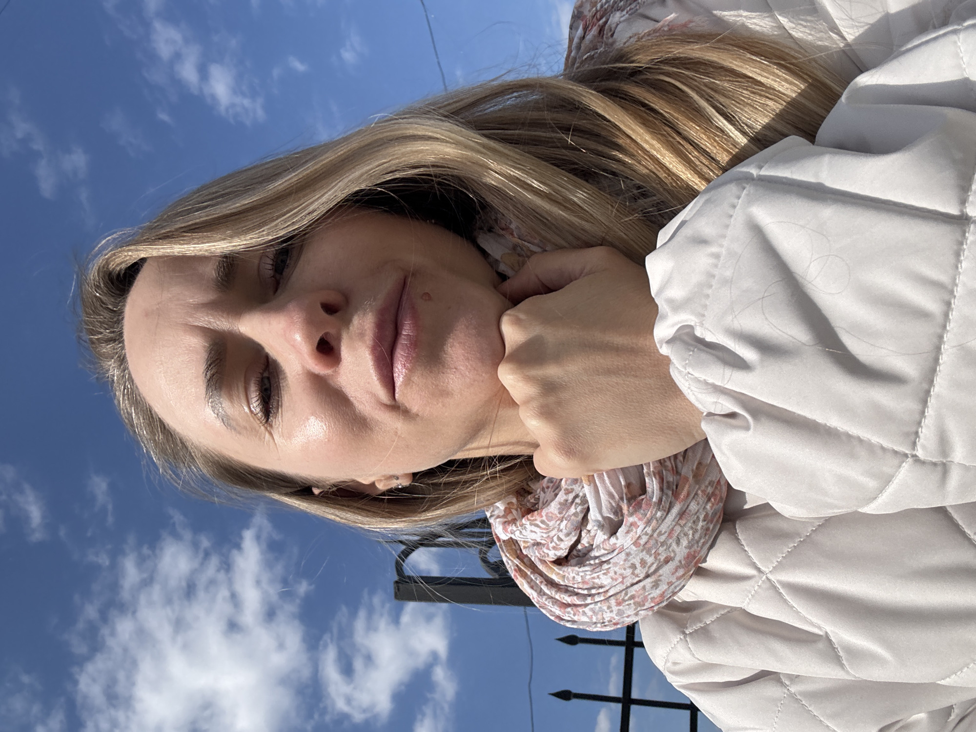 This media file shows a self-portrait of a woman taken outdoors, from a low-angle perspective, with the subject looking upwards towards the camera. The woman, appearing to be in her 20s or 30s, has long, light-colored hair and is illuminated by bright sunlight, casting highlights on her skin and hair. Her right hand is resting gently on her chin or jawline.
She is wearing a light-colored, possibly off-white or cream, quilted jacket. Around her neck, a patterned scarf with shades of pink, cream, and potentially light brown is visible. The background consists primarily of a clear, bright blue sky with scattered white, wispy clouds, indicating a sunny day. A dark metal fence with pointed top finials is partially visible in the bottom right corner, suggesting the location is adjacent to a public or private outdoor area. The woman's expression is calm, displaying a subtle smirk or small smile. The image is taken in bright daylight. No visible text is present. The provided context indicates the location is Chaikovsky, Russia, though no specific landmarks are visible in the image itself.