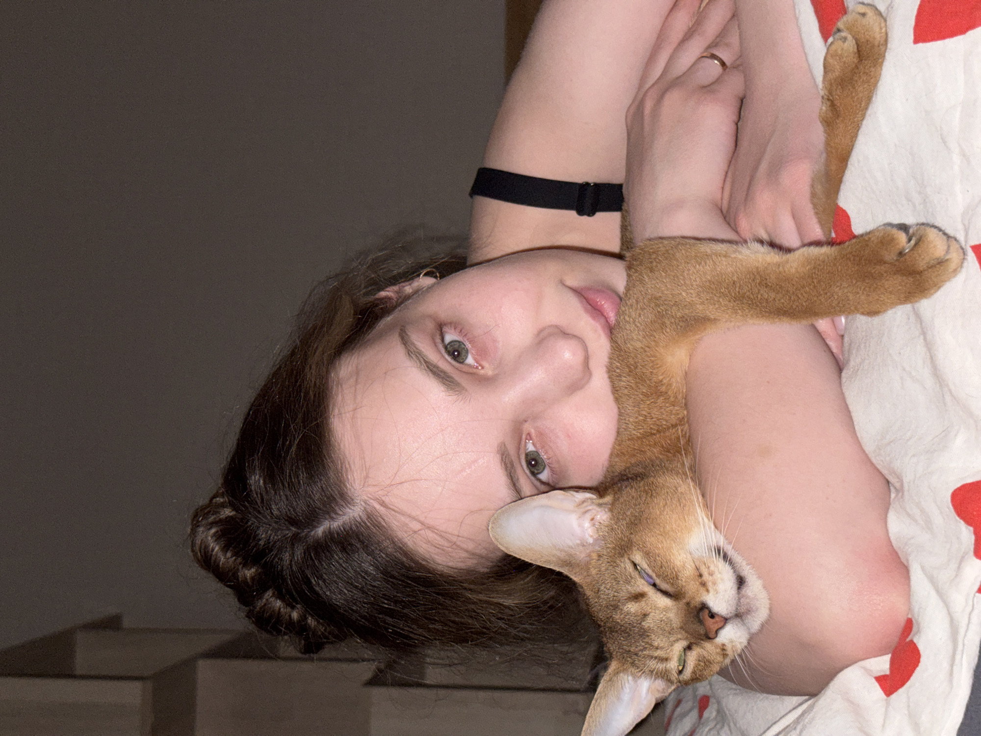 The image features a young woman holding a golden-brown cat in her arms. She is lying on her side, with the cat nestled against her arm. The woman has light skin, green eyes, and brown hair partially braided. She is wearing a black choker. Her arms are crossed in front of her. A gold ring is visible on her left hand. The cat has one eye slightly open and a light pink nose. It is lying against the woman's arm, appearing relaxed. They are lying on a white blanket with red heart patterns. The background is dimly lit, with a grey wall and what appear to be stairs partially visible. The location is described as the city of Chaikovsky, Russia.