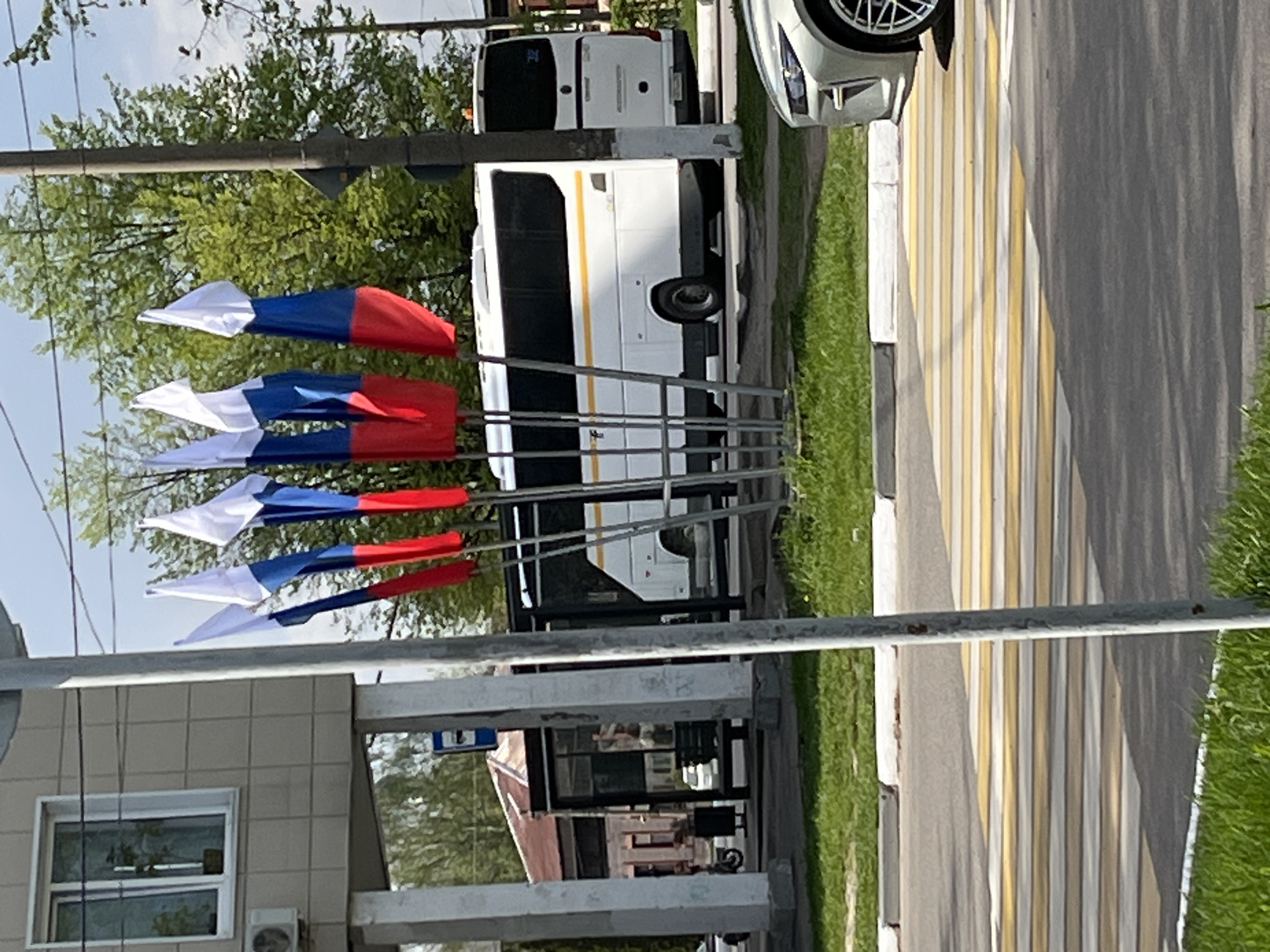 The image depicts a street scene, potentially in Noginsk, Russia, based on the presence of flags with the white-blue-red color scheme of the Russian flag. There are several small flags with this color scheme mounted on vertical poles. A white minibus with a yellow stripe is parked near the flags. A pedestrian crossing with alternating white and yellow stripes is visible on the street. The environment includes green grass, trees, and buildings. One building appears to be a one-story structure with a window and an air conditioning unit. A bus stop is near the flags. A portion of a white car is visible at the top of the image.