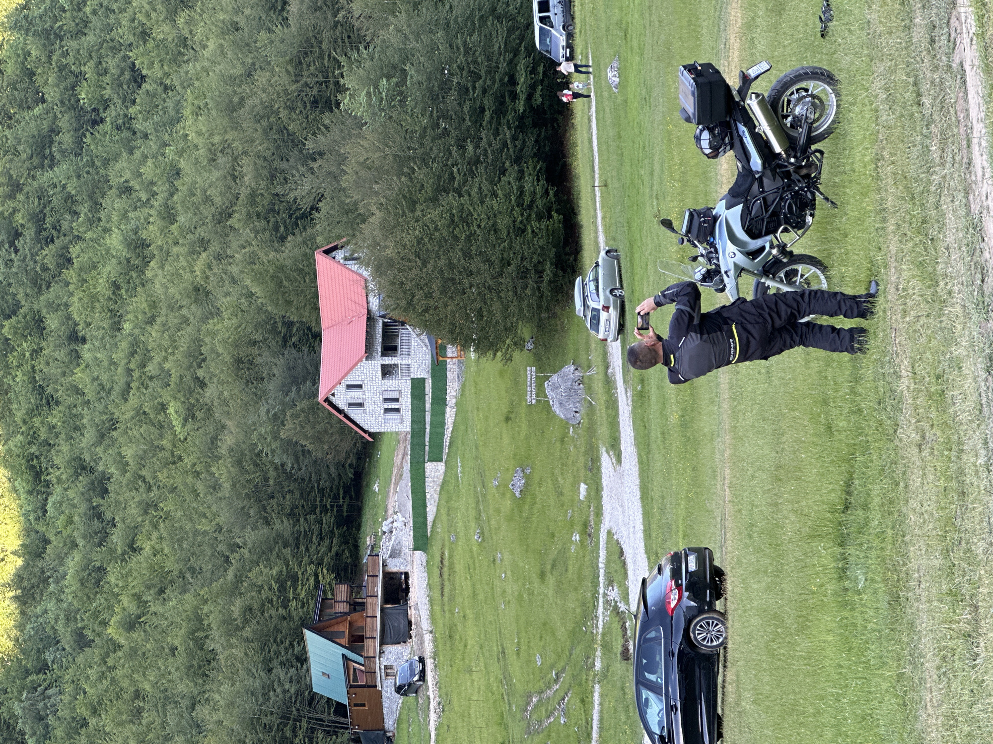 In the city of Dolja, Montenegro, a man in a black and yellow motorcycle suit stands in a grassy field, holding up a smartphone to take a picture of a light blue motorcycle parked in front of him. The motorcycle is equipped with saddlebags and a top case. In the background, several houses and trees are visible, with a dense forest covering the hillside. A black car is parked to the left of the frame, and another silver car is visible further up the hill, near a group of people walking. A van is parked on a dirt road in the distance. The scene appears to be set in a rural, mountainous area.