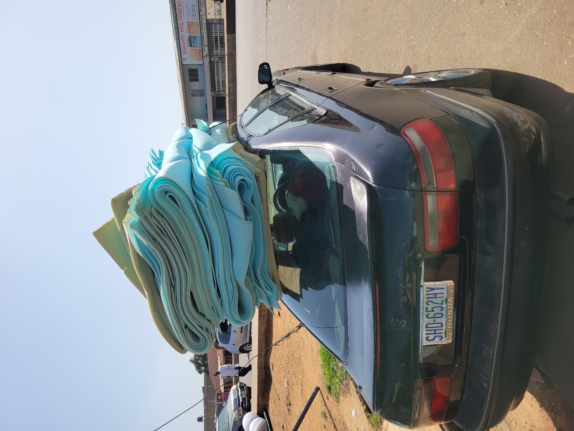 The image depicts a black Honda car precariously loaded with a large stack of blue and green foam sheets. The car's license plate reads "SHD-652HY." The scene appears to be on a street, with buildings visible in the background, including one displaying signage for "SOARMIC MULTIMEDIA." There are other vehicles and a few individuals visible in the background as well. The location is in the city of Unknown, Unknown.