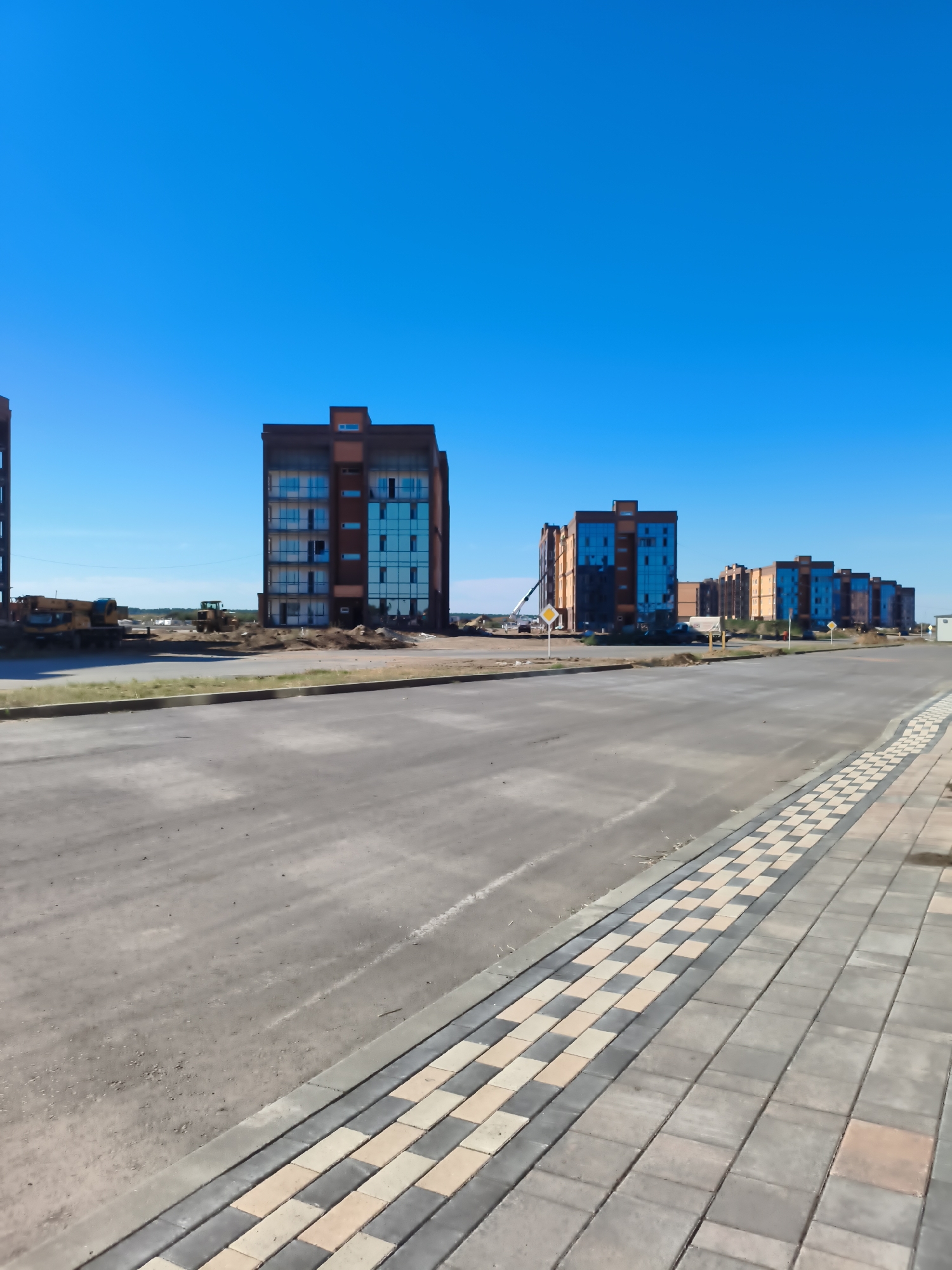 This is a medium-wide, eye-level, full shot of a construction site in Мичурин ауылдық округі, Kazakhstan. The foreground features an asphalt road with a decorative paved walkway on the right, composed of grey, beige, and orange rectangular stones. The walkway is bordered by a concrete curb. In the mid-ground, several multi-story apartment buildings, some with modern glass facades and brown exteriors, are under construction. Construction equipment, including yellow excavators, are visible on the dirt and gravel ground surrounding the buildings. The background is dominated by a clear, bright blue sky.