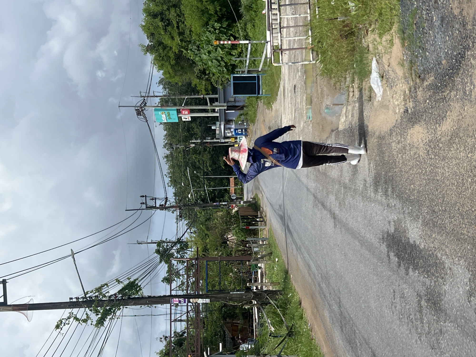 A person wearing a blue hooded jacket, black pants with white stripes, and white boots walks away from the camera on a paved road in Pa Khlok, Thailand. The individual is also wearing a wide-brimmed hat. The road is lined with vegetation and utility poles carrying numerous wires. In the background, there is a blue sign with a white "Lotus" logo and text, and behind that, a building and more trees under a cloudy sky.