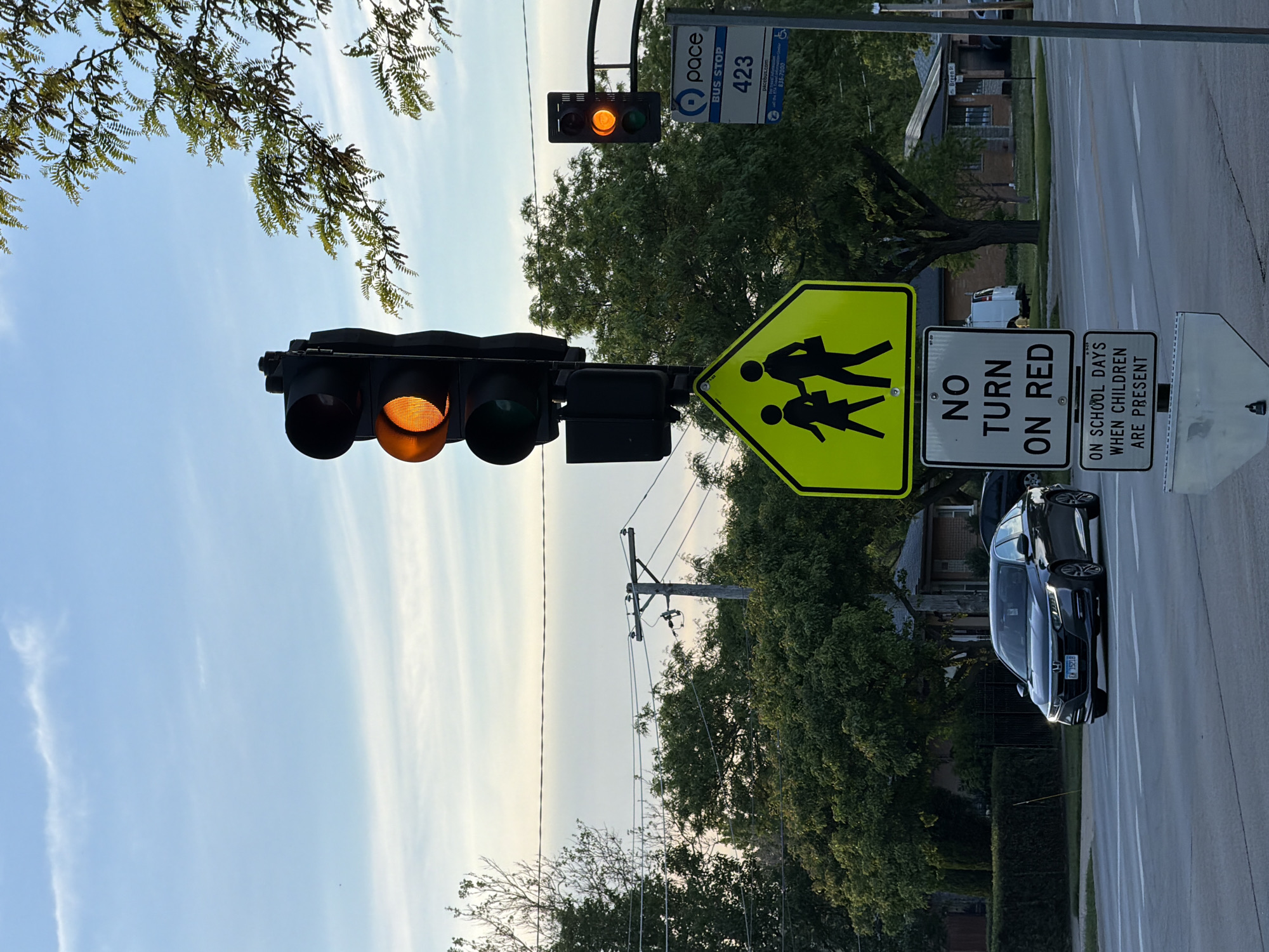 The image captures an intersection in the city of Niles, United States of America. The traffic light is yellow. Adjacent to the traffic light are signs indicating a school zone and restrictions on right turns on red when children are present. A PACE bus stop sign is also visible. In the foreground, a black car is stopped at the intersection. The surrounding environment includes trees, residential buildings, and utility poles.
