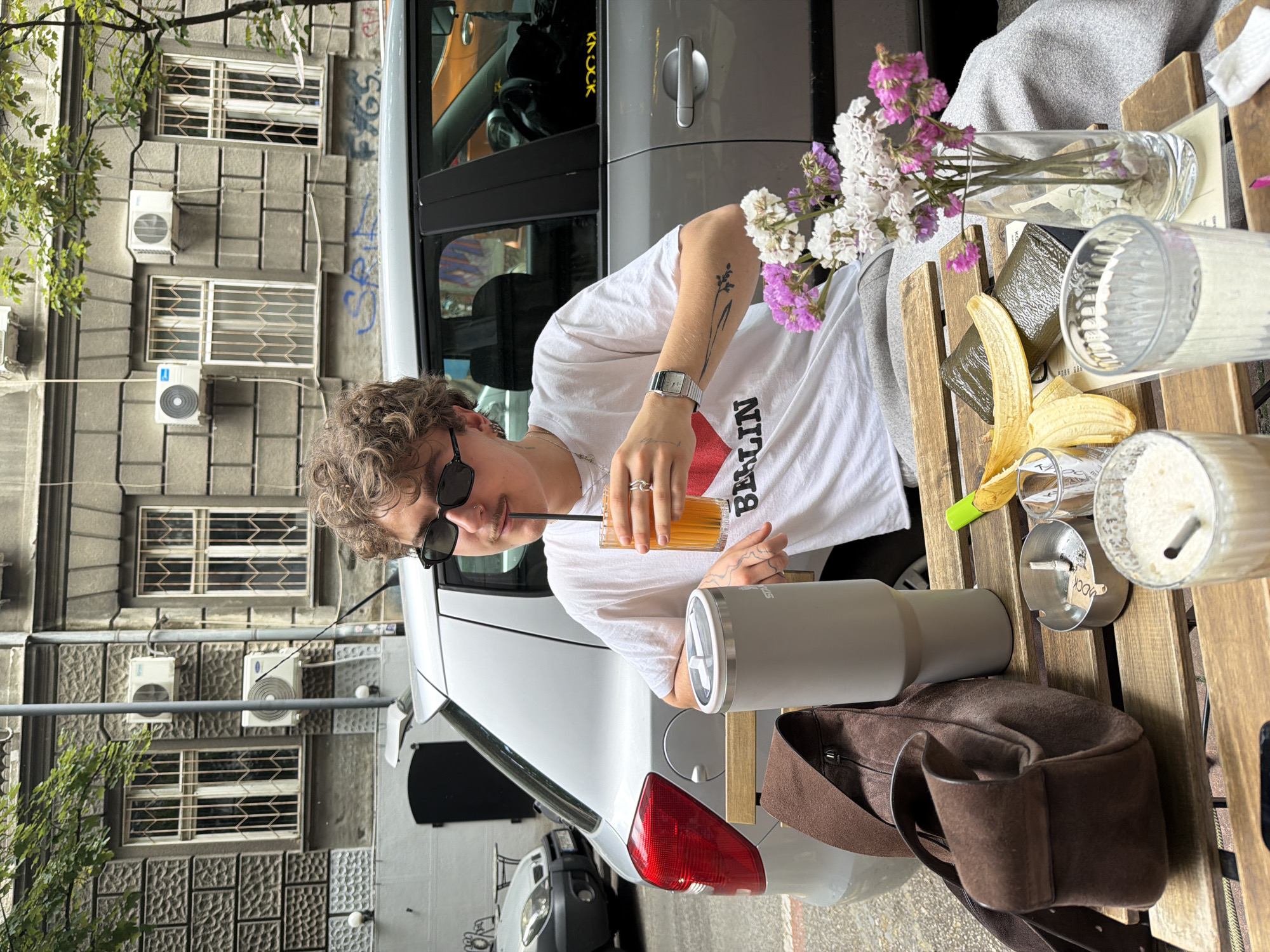 A person with curly blonde hair and sunglasses sits at an outdoor table. The person wears a white T-shirt with "Berlin" printed on it, a silver watch, and sips from a tall glass with a straw. A partially peeled banana, a vase of flowers, an ashtray, a large gray drinking cup, and a brown suede bag are on the table. In the background are cars and a building with numerous windows and air conditioning units. The location is Belgrade, Serbia, based on "Mock" sign on the ashtray.