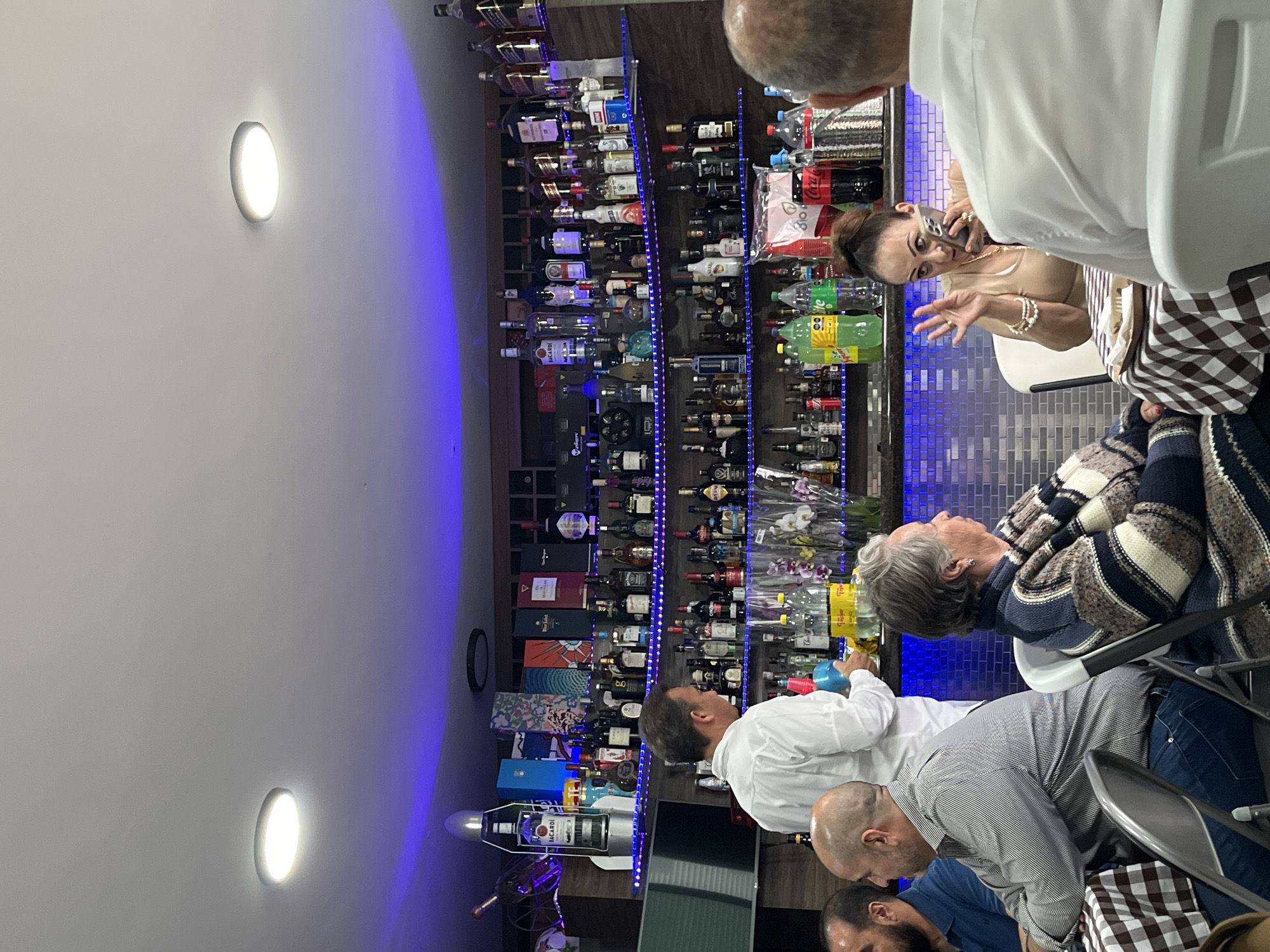 The scene depicts a gathering of people inside what appears to be a bar or restaurant. A woman is talking on a phone while seated at a table. There is a well-stocked bar with a variety of liquor bottles and other beverages behind her. Several other people are seated around the table engaged in conversation. The interior lighting includes recessed lights in the ceiling and blue accent lighting highlighting the bar area. The location is identified as San Bernardino Tlaxcalancingo, Mexico.