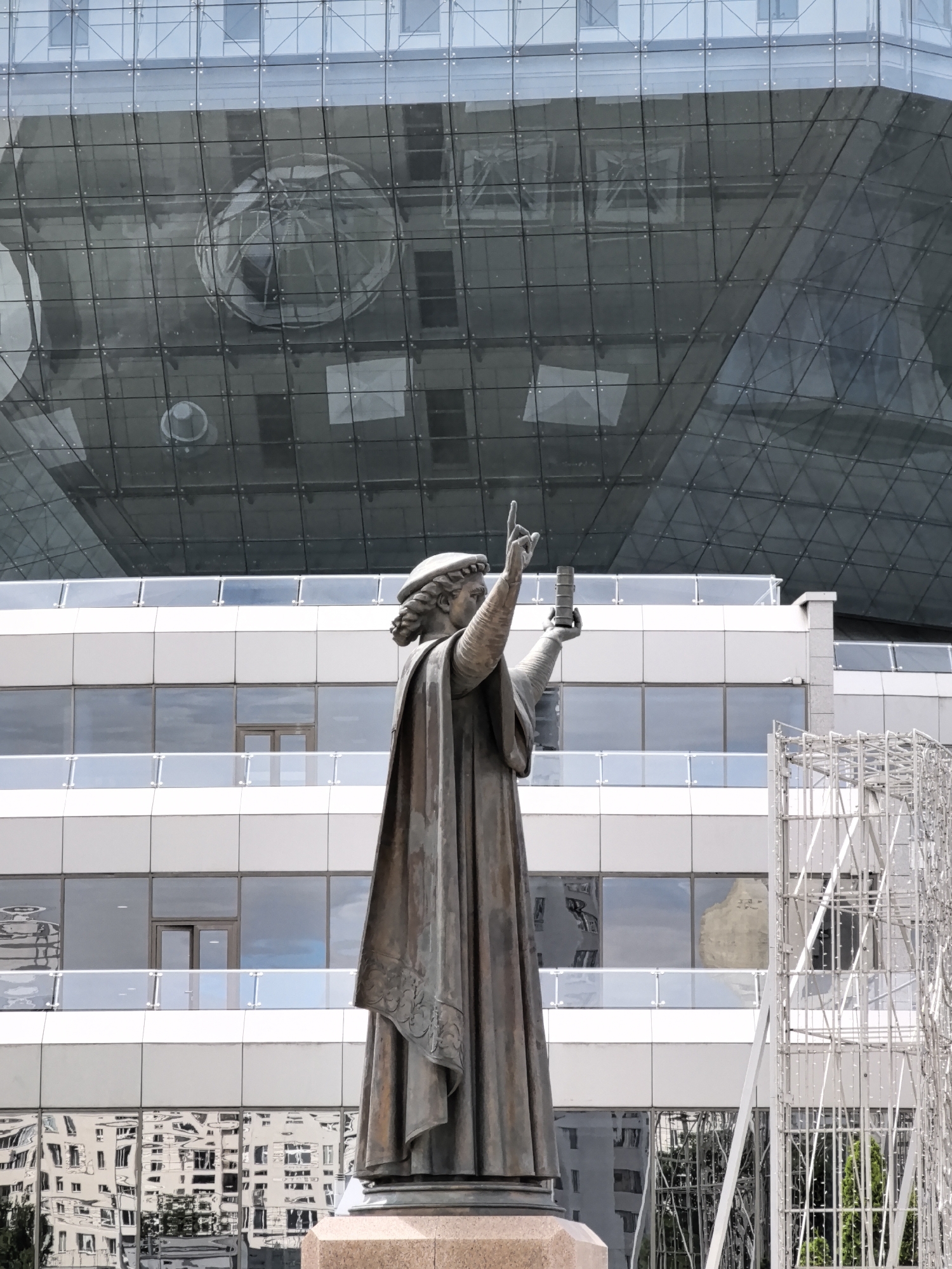 A bronze statue of a historical figure stands in front of a modern building. The statue depicts a man wearing a cap and a long, flowing robe, holding a cylindrical object in his left hand and pointing upwards with his right index finger. The building behind the statue is a large, multi-story structure with a facade primarily composed of glass panels and white composite materials. Reflections of other buildings and the sky are visible on the glass surfaces. A metal scaffolding structure is partially visible to the right of the statue. The scene appears to be an outdoor public space.