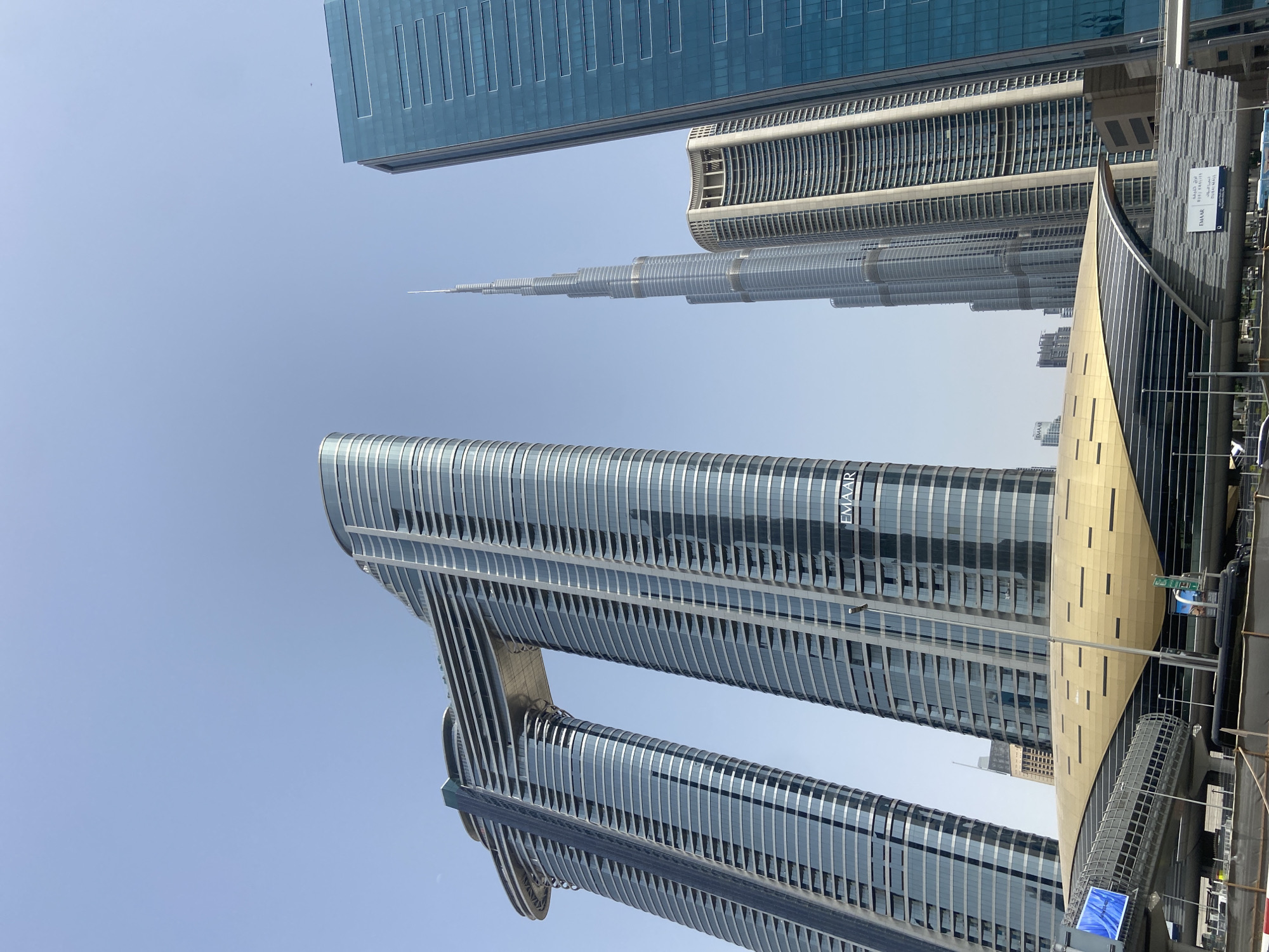 The image shows a view of several skyscrapers in Dubai, United Arab Emirates, under a clear, light blue sky. In the foreground is a pair of connected skyscrapers with a distinctive bridge structure, marked with "EMAAR" signage on the upper portion. Behind these is the Burj Khalifa, easily recognizable by its unique tapering shape, and a neighboring high-rise building. To the right of the image, a partially visible modern structure with golden panels suggests a public transit station or similar infrastructure. The buildings feature reflective glass facades, indicating a contemporary architectural style. No people are visible.