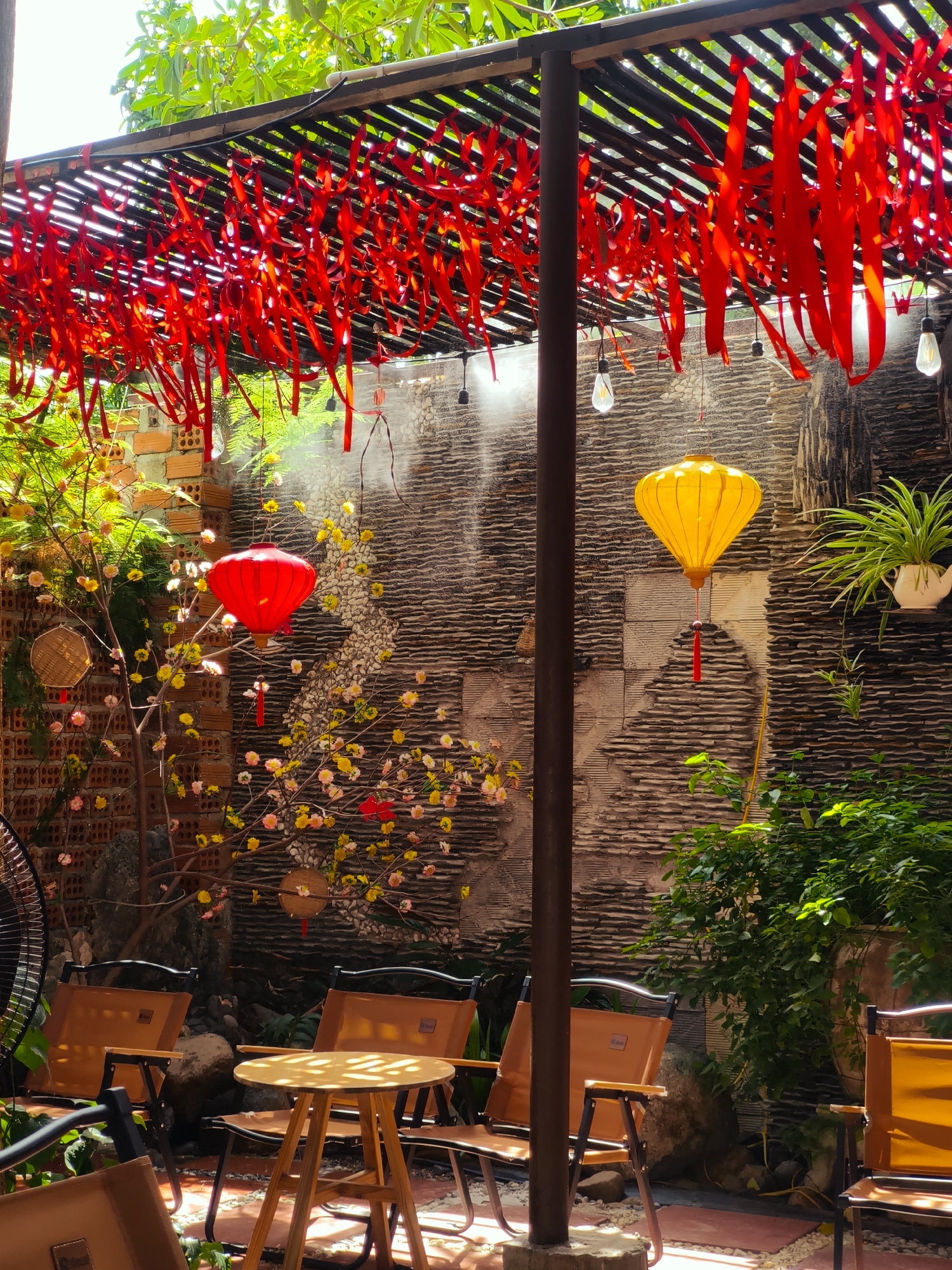 This image depicts an outdoor seating area, likely a cafe or restaurant, in Cam Ranh, Vietnam. The scene is decorated with numerous red streamers hanging from a slatted roof, creating a festive atmosphere. Two lanterns are visible: a red one on the left, hanging from a branch with small yellow and pink flowers, and a yellow one on the right, suspended from the roof structure. The wall in the background is textured with horizontal stone slabs. Several brown camping-style chairs are arranged around a small wooden table, with a fan visible in the lower left corner. Lush green foliage and brick walls are also present in the background, indicating an outdoor, possibly garden setting. The lighting suggests it is daytime, with sunlight filtering through the roof and foliage.