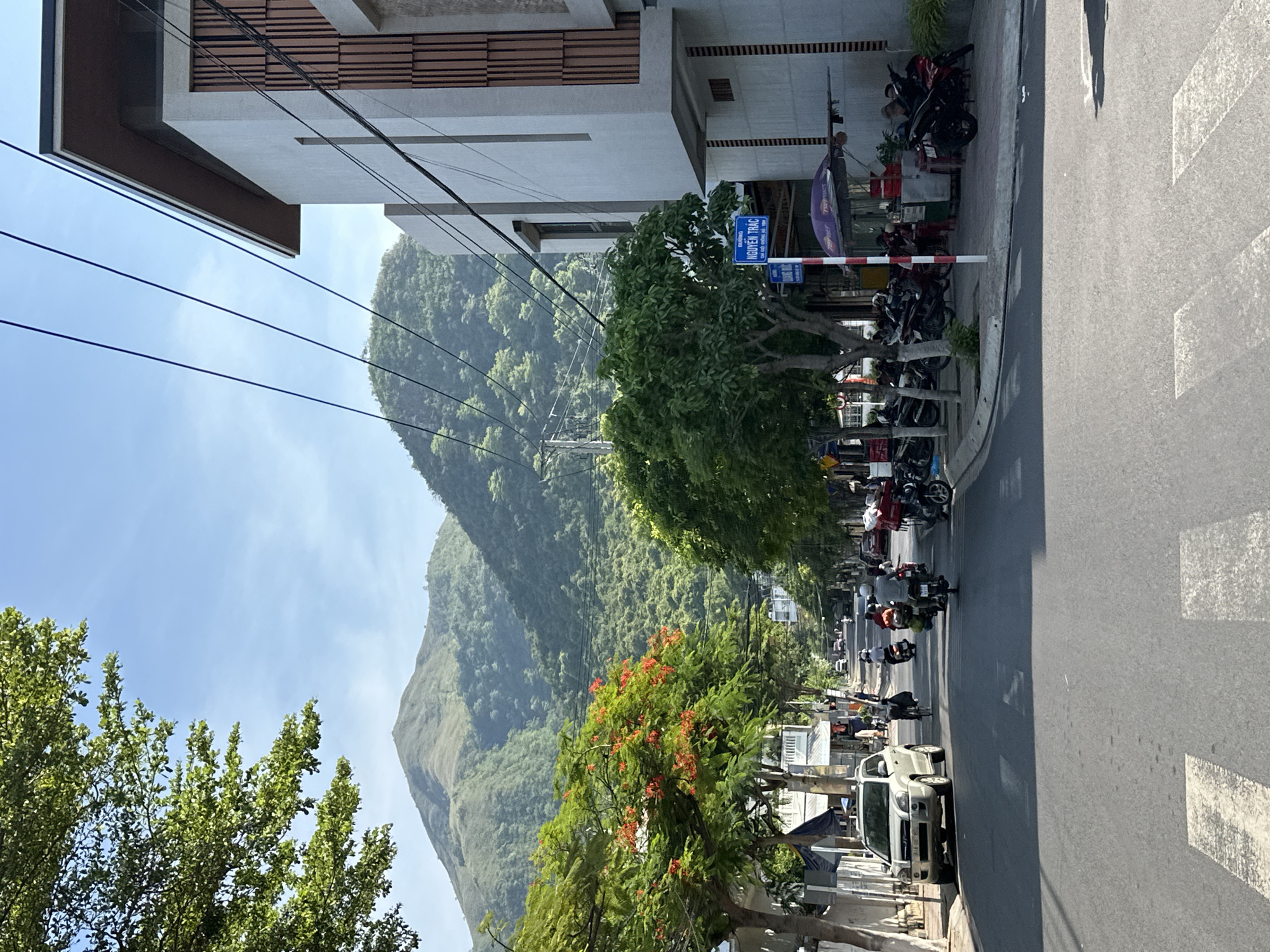 The image depicts a street scene in Bac Nha Trang Ward, Vietnam. In the foreground, a paved road with white pedestrian crossing lines is visible. The road is lined with trees and buildings on either side. A silver SUV is parked on the left side of the road, and several motorbikes are visible on the road and parked on the sidewalk.
The left side of the street features a row of low-rise buildings and trees, one of which has vibrant orange flowers. A large, lush green tree with small leaves is prominent in the mid-ground on the left.
The right side of the street has a modern, multi-story building with a light-colored facade and wooden slats. A blue street sign with white text reading "NGUYEN TRAC" is attached to a pole on the sidewalk. A small kiosk with a purple umbrella is situated on the sidewalk in front of the building.
In the background, a large, verdant mountain range dominates the skyline, with its slopes covered in dense green vegetation. The sky is clear and blue. The scene appears to be captured during daylight hours, with sunlight casting shadows on the road. Several people are visible, primarily riding or tending to motorbikes on the street and sidewalk. The overall atmosphere suggests a busy, tropical urban environment with natural landscapes as a backdrop.