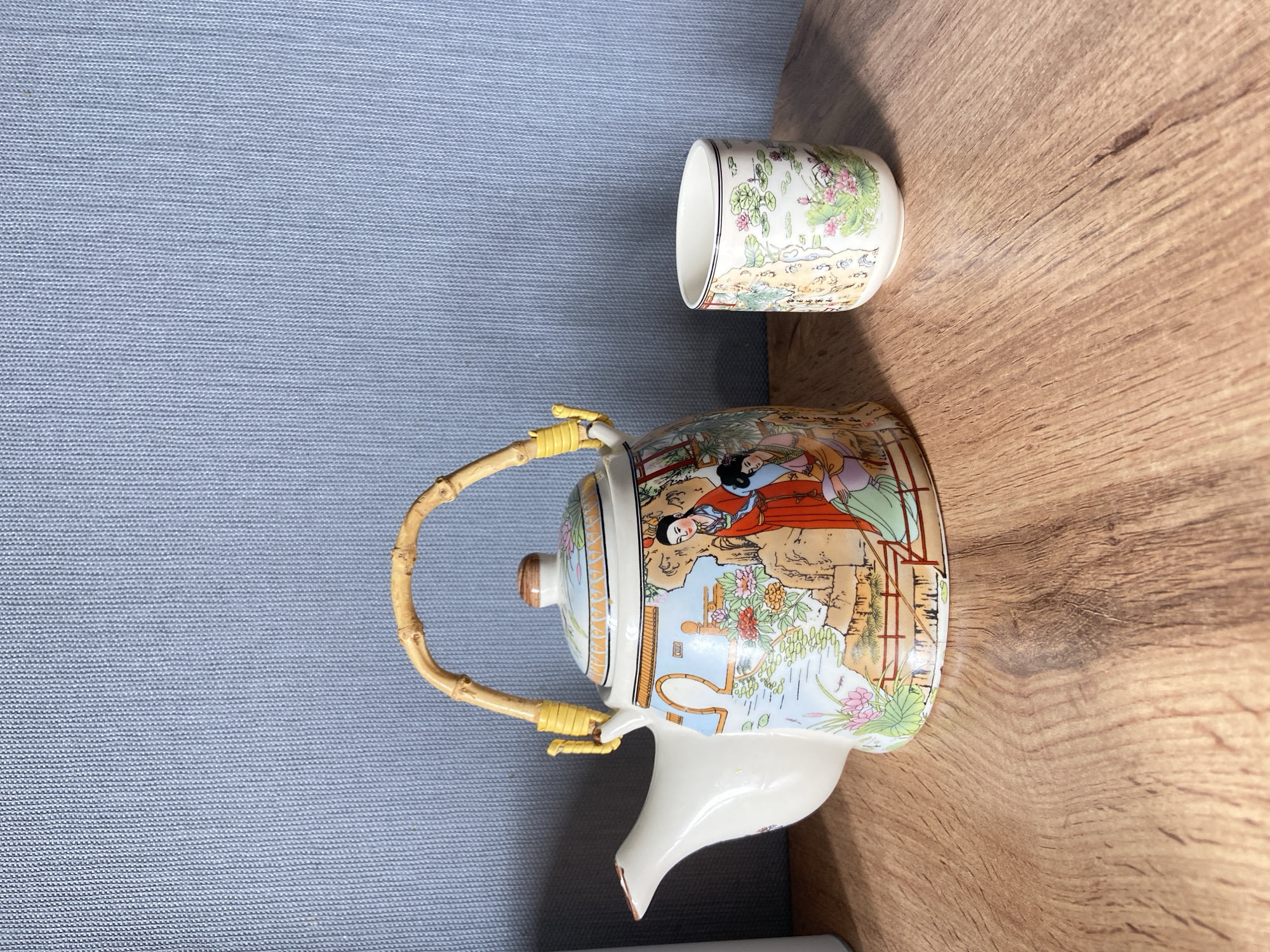 A teapot and cup set, decorated with traditional Chinese artwork featuring women in gardens and nature scenes, sits on a wooden table against a blue textured background. The teapot has a bamboo handle and yellow accents. The cup is smaller and cylindrical, with a similar floral design. The lighting suggests an indoor setting, likely a home or shop. The scene is static and depicts decorative tableware.