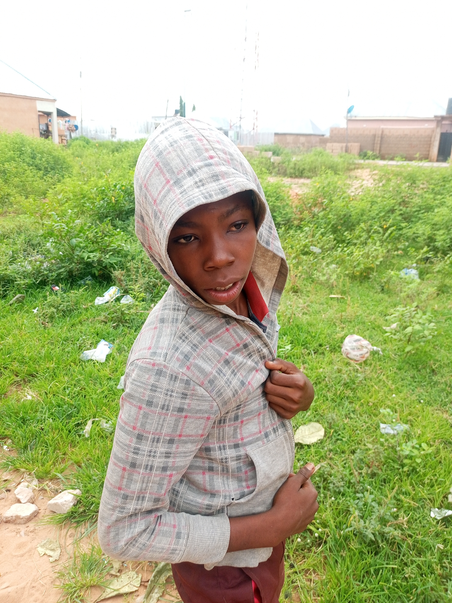 A young person wearing a grey hooded sweatshirt with a plaid pattern is standing in a grassy area with some debris. The person is facing the camera from the right side of the frame, with their head tilted slightly. The background shows some buildings and vegetation, consistent with a location in Tempure, Nigeria. The person's left hand is holding the front of their sweatshirt, and their right hand is resting on their stomach. The grass is green and appears to be somewhat unkempt, with several pieces of white and colored plastic waste scattered throughout. The sky is overcast and bright.