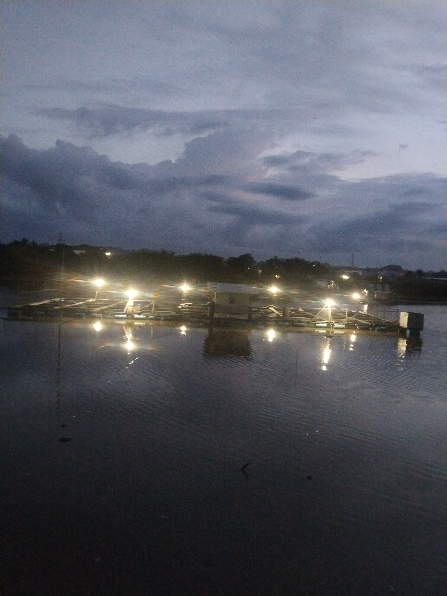 The image depicts a body of water at dusk or night, likely located in Lhokseumawe, Indonesia. A floating structure with several illuminated lights is situated in the water. The structure appears to be a rectangular platform or raft, possibly used for aquaculture or other activities. A small, enclosed building is also visible on the platform. The water reflects the lights, creating streaks across the surface. The sky is overcast with dark clouds. In the background, there are trees and hints of a built-up area, suggesting the presence of the city. There are no people visible in the image.