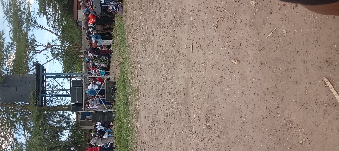 The image shows a dirt field in Kitengela, Kenya, with a water tower and a gathering of people. The water tower is a cylindrical structure with a blue metal frame. Behind the water tower are large speakers and a small, light-colored structure. Several people are gathered in front of the water tower, some sitting and some standing. The people are wearing various casual clothing. Some of the people have their hands raised, while others are interacting with each other. The environment is dry and dusty, with scattered grass and leaves. The sky is blue and cloudless. The image was taken from a low angle, looking up at the water tower and the people. The ground is mostly dry and dusty, with a few patches of grass. The image is taken in front of the water tower, where people are gathered. The field appears to be used for various purposes, including events and gatherings.